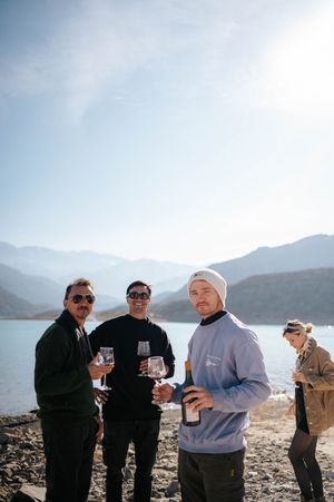 Three men holding wine glasses and bottles by a mountain lake on a clear day.