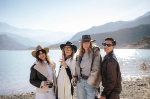 Four women posing with wine glasses near a mountain lake wearing hats and sunglasses.