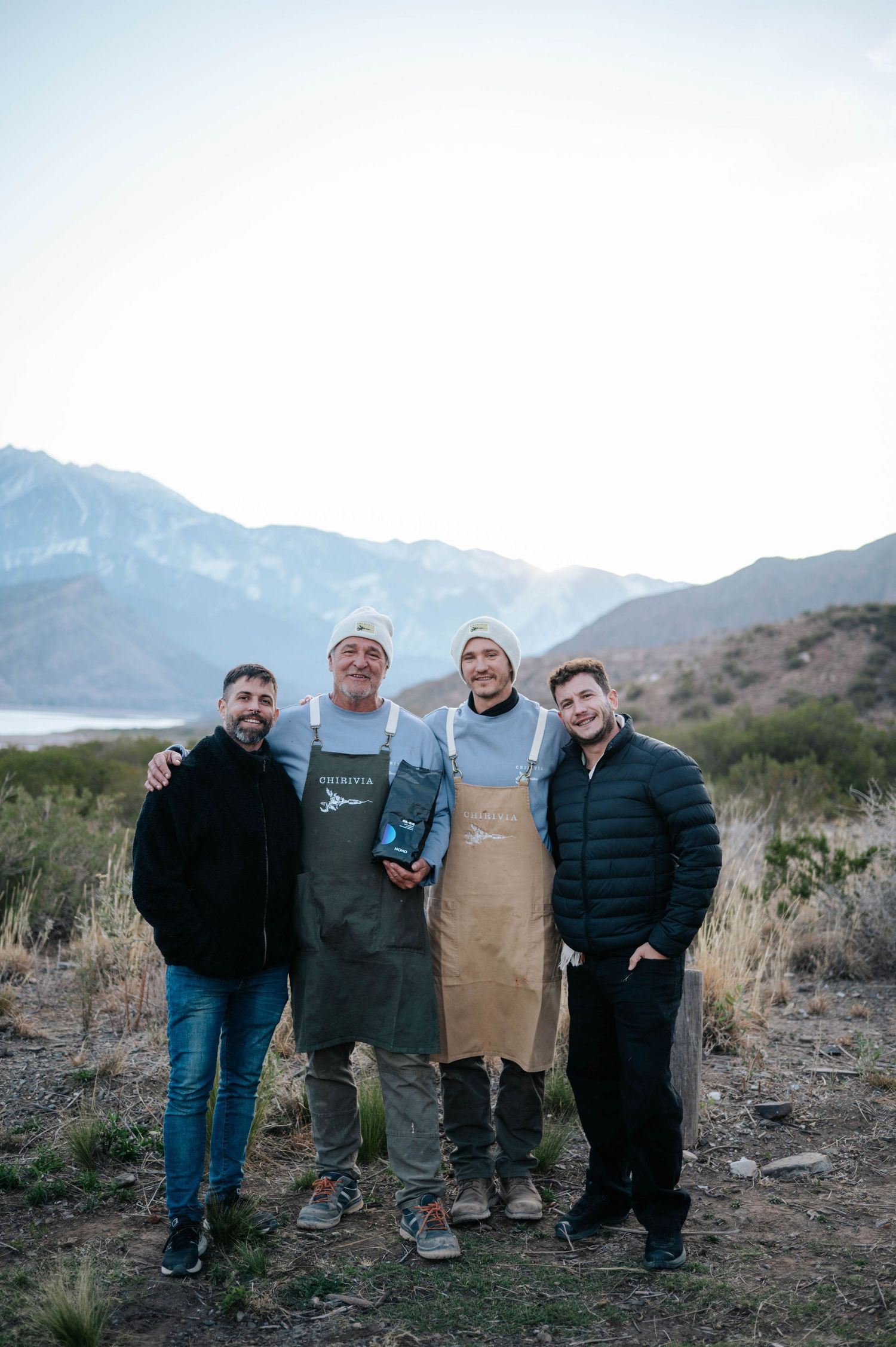 Four men posing in aprons near a mountain lake at an outdoor culinary gathering.