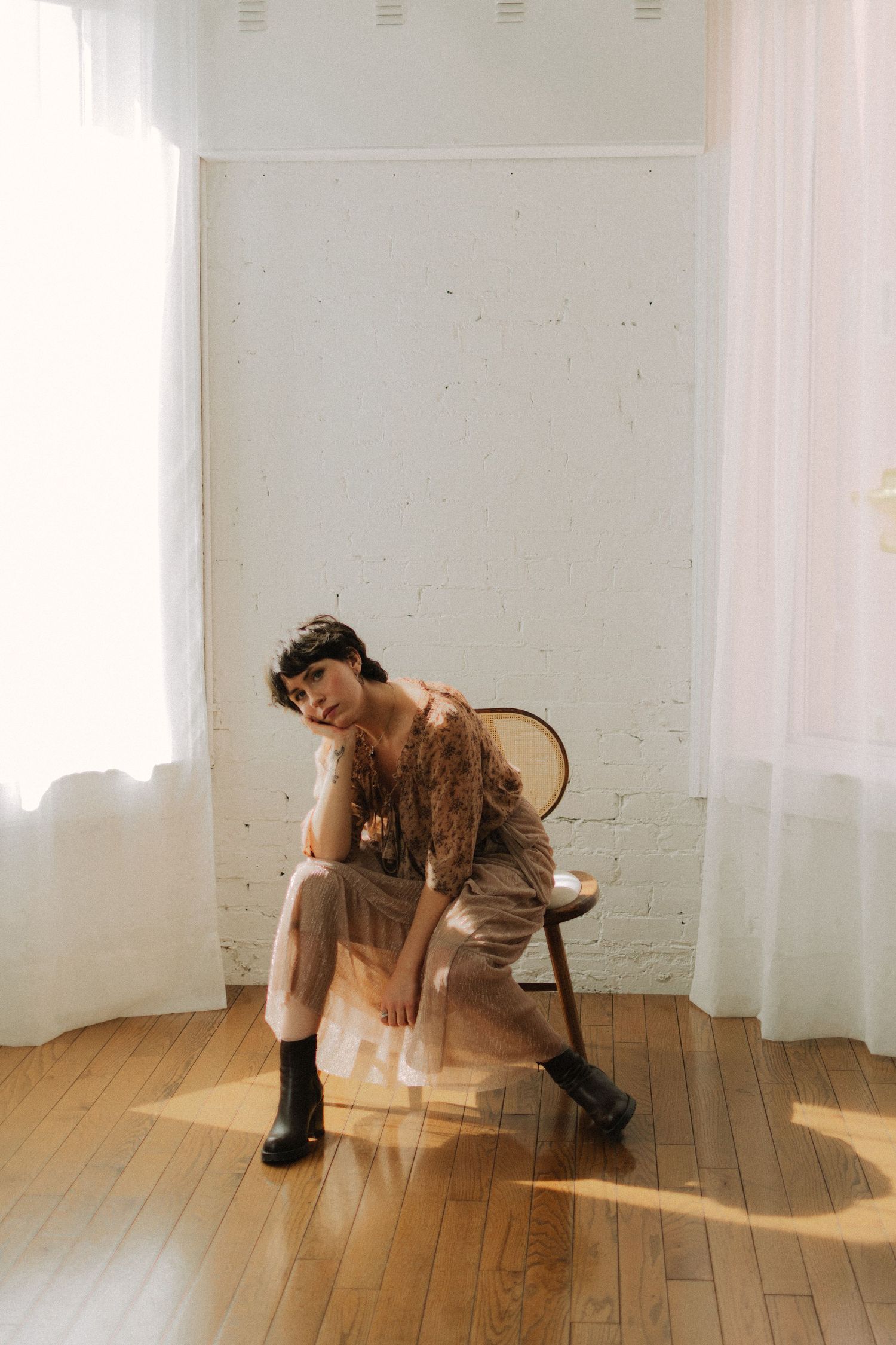 Figure in a vintage dress and boots sits pensively on a wooden chair in a sunlit room with white curtains.