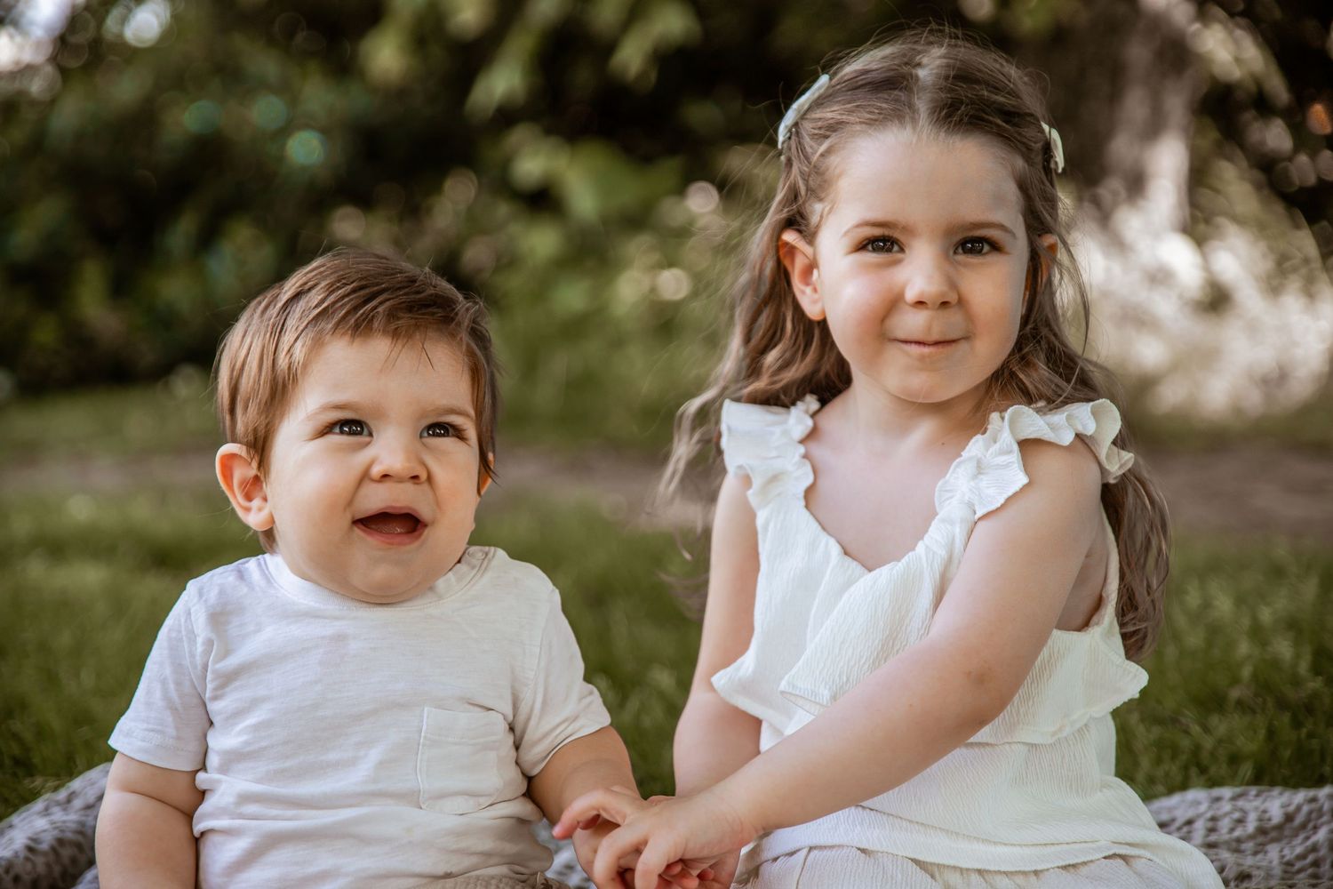 Two young children in white outfits sit together outdoors during a sunny portrait session.