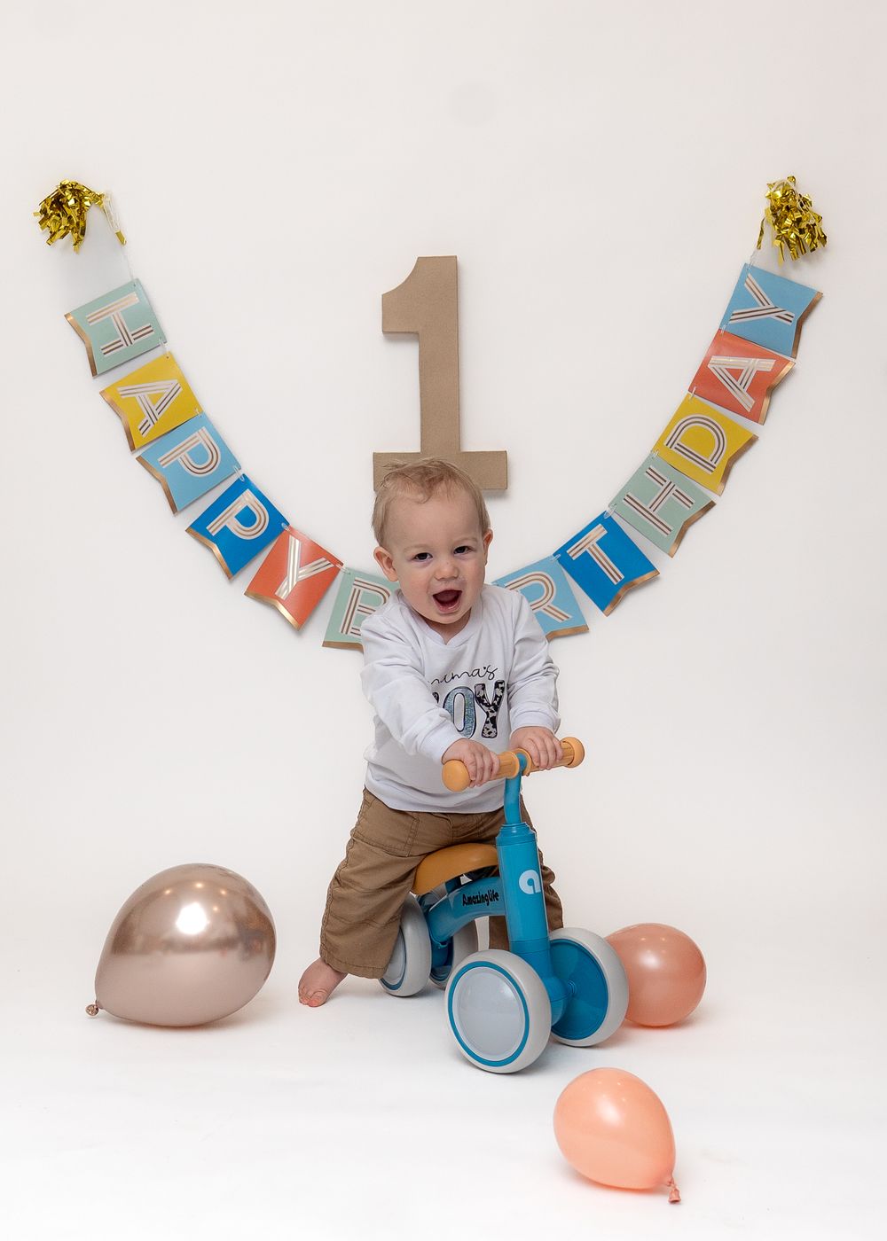 Child celebrating first birthday with happy expression on blue balance bike surrounded by balloons and birthday banner.