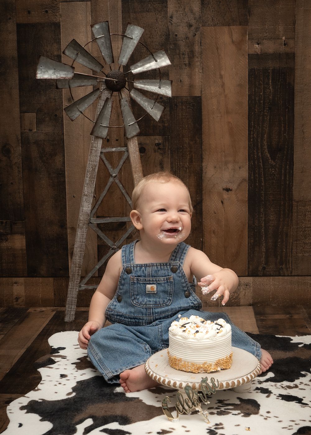 Smiling baby in denim overalls sitting with a cake near rustic windmill backdrop and cowhide rug.