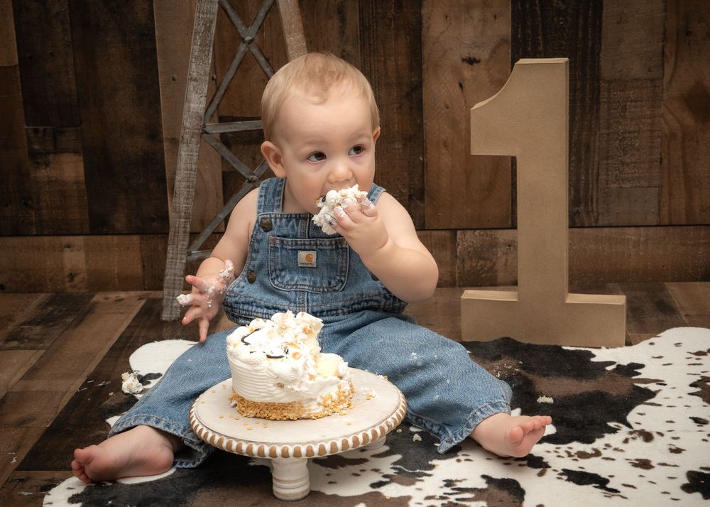 Baby in denim overalls sits on cowhide rug with birthday cake on vintage pedestal stand during cake smash photoshoot.