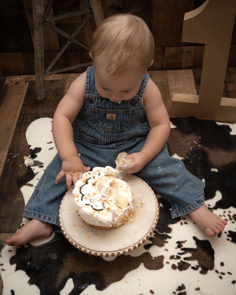 Small child in overalls sits on a cowhide rug while reaching for a cake on a white decorative plate.