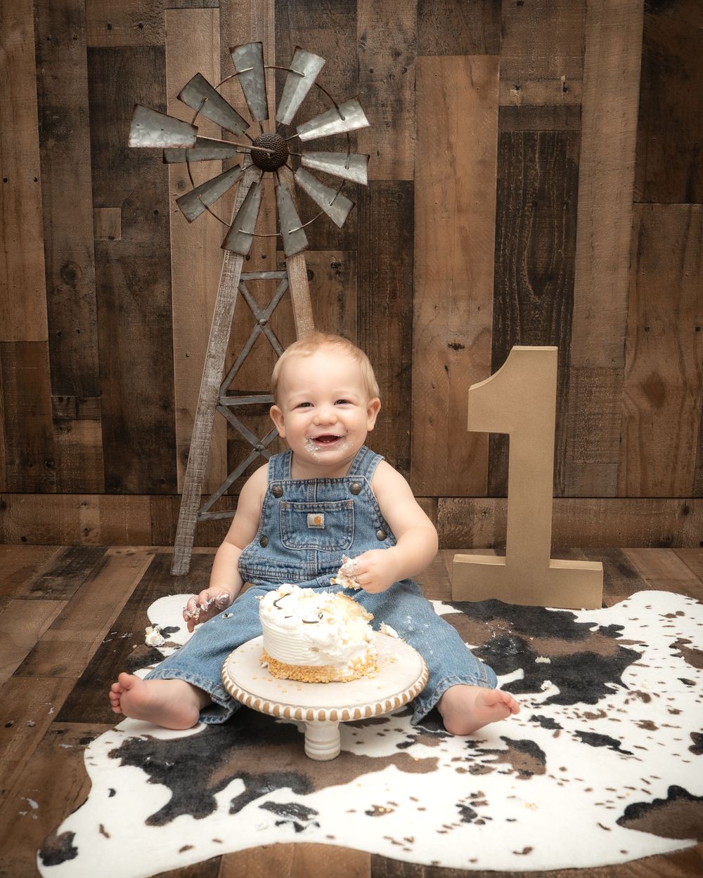 A rustic farmhouse scene with denim overalls, wooden walls, decorative windmill and cow print rug for a first birthday celebration.