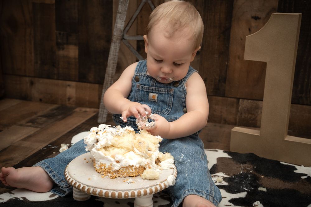 Small child in overalls doing cake smash with white frosted cake on rustic wooden background.