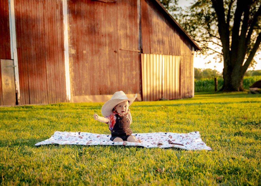 A small figure sits on a blanket in front of a rustic red barn during golden hour.
