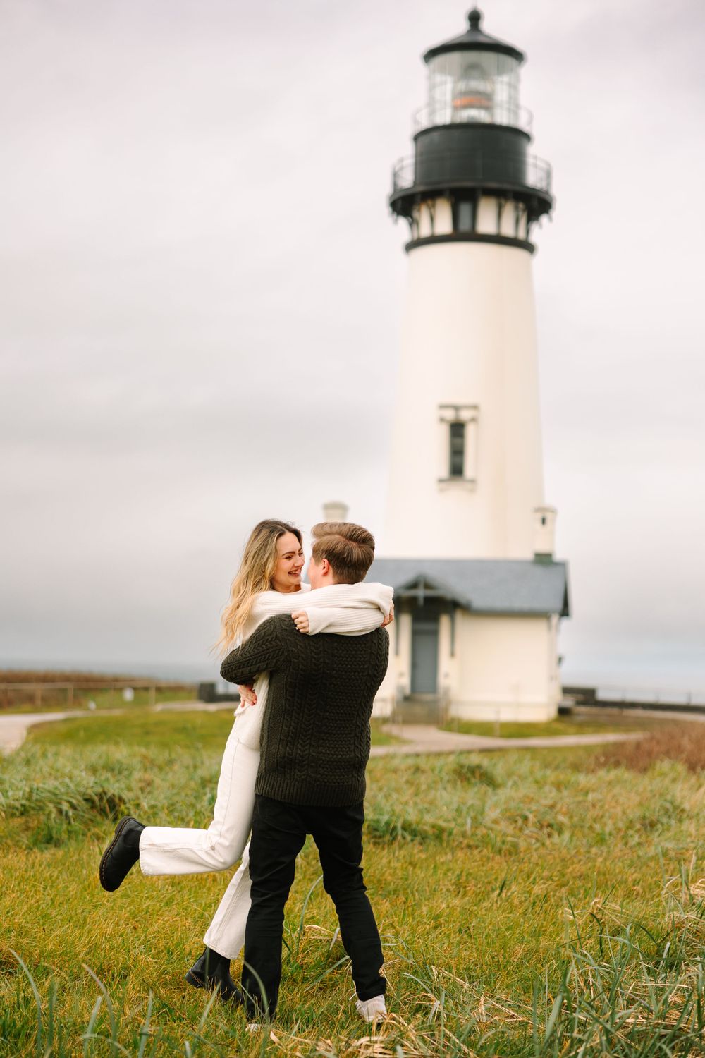 Romantic moment in front of classic white lighthouse on grassy coastal cliff.