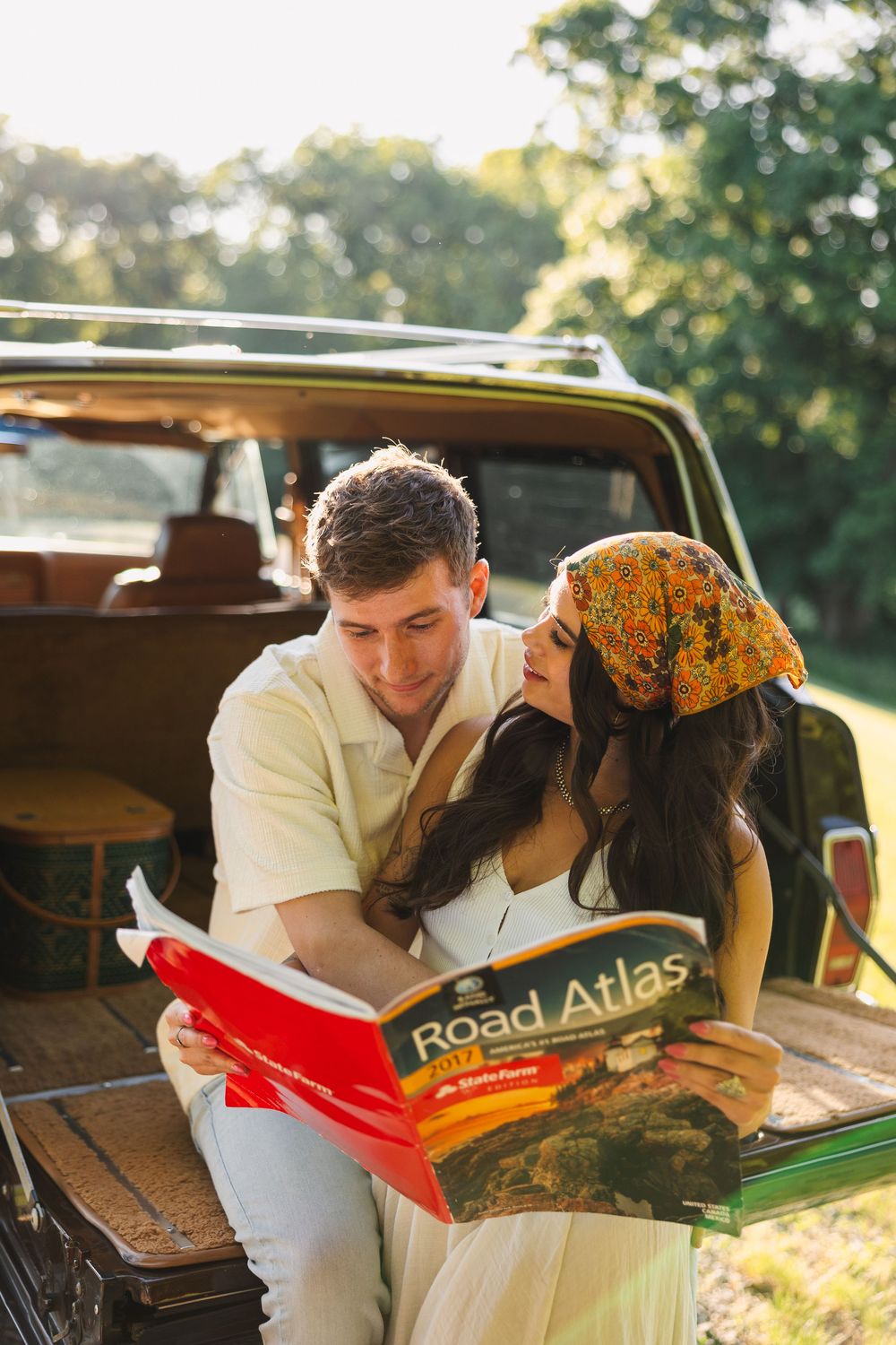 Couple sitting on vintage car trunk reading a Road Atlas together outdoors in golden sunlight.