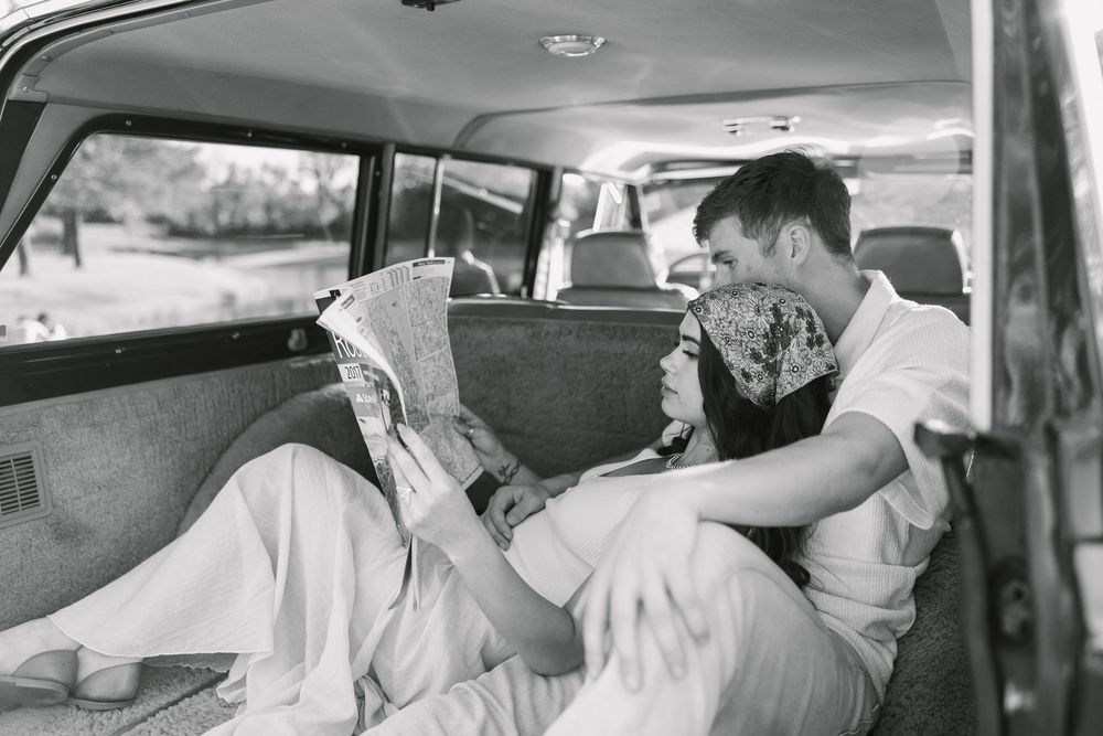 Couple relaxing in vintage car backseat, sharing newspaper in black and white romantic photo.