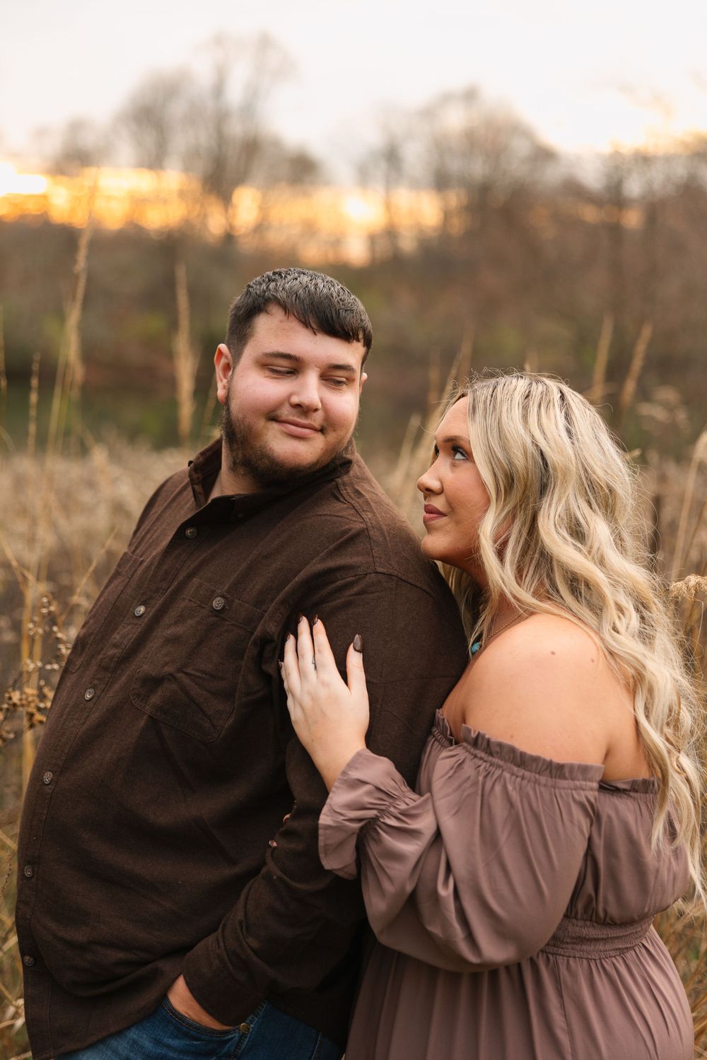 A couple poses together in a field during golden hour wearing brown and mauve tones.