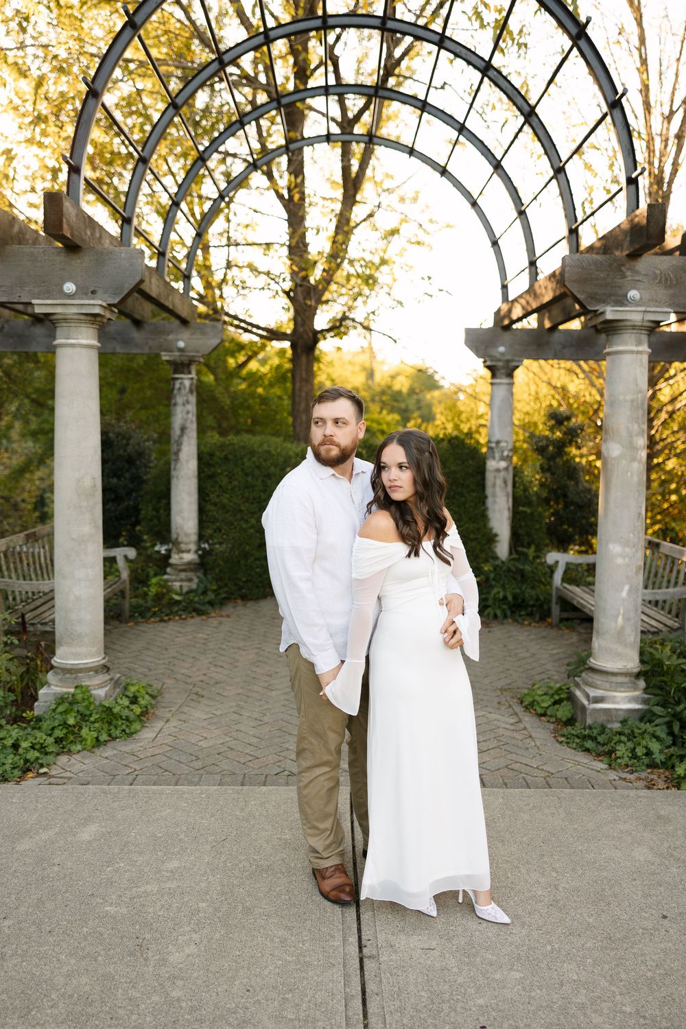 A couple in white outfits poses together in a romantic garden setting with stone columns and autumn foliage.