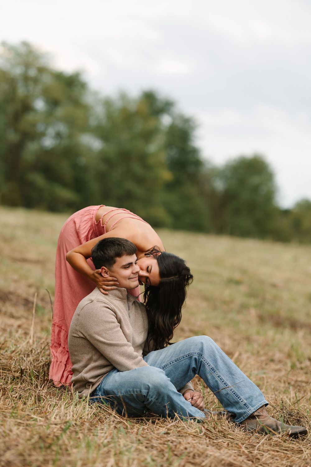 Couple embraces while sitting in a grassy field on an overcast day.