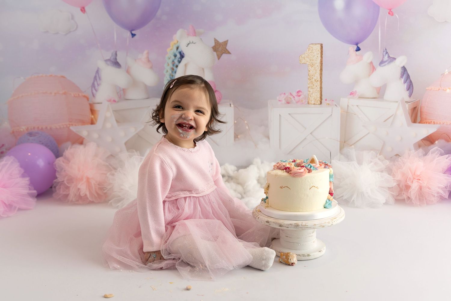 First birthday celebration setup with purple balloons, pink tulle decorations, and white cake on gold stand.