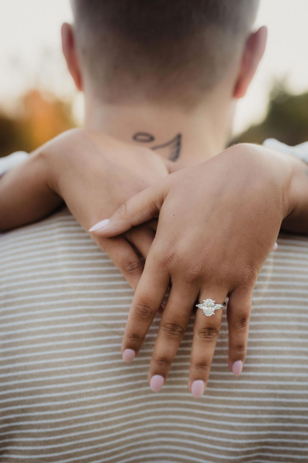 Close-up of an engagement ring on a hand resting against a striped shirt with a neck tattoo visible.