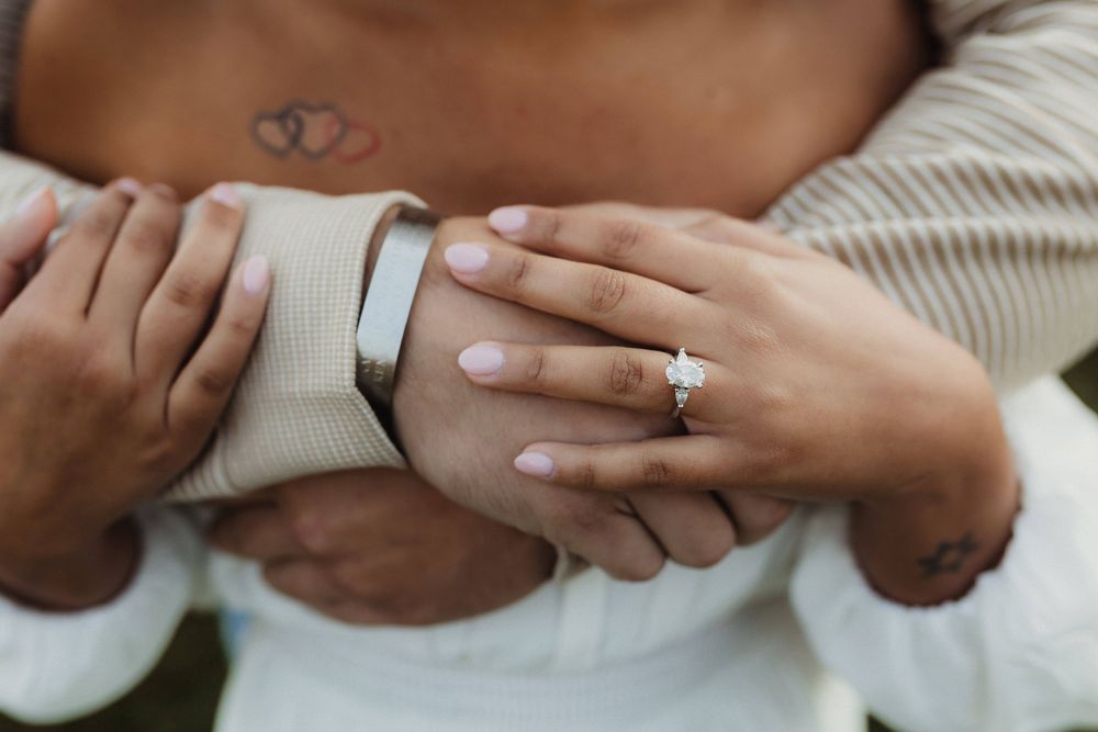 Close-up of hands with engagement rings and heart tattoo against white clothing.