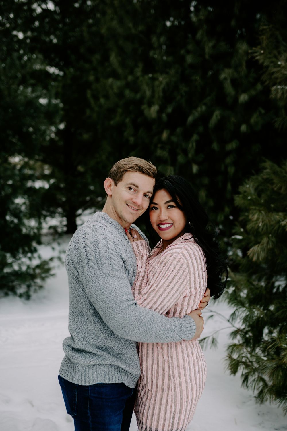 Winter engagement photo of couple embracing in knit sweaters against snowy evergreen backdrop.