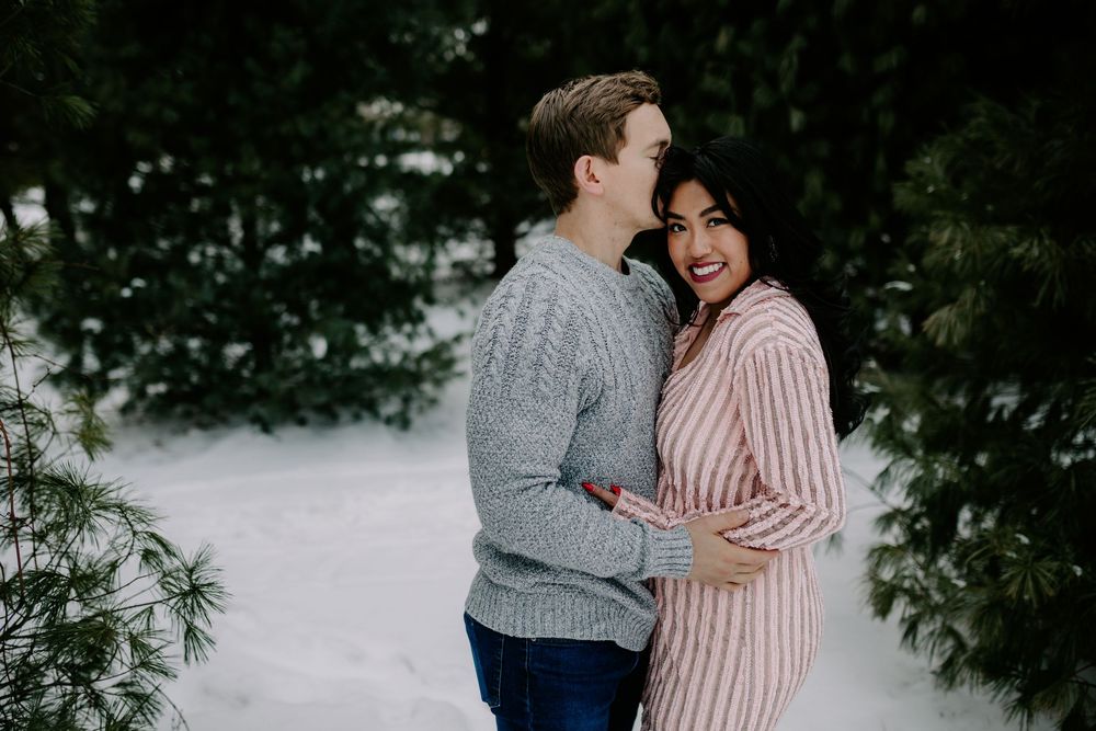 A couple embraces in a romantic winter scene with snow and evergreen trees in the background.
