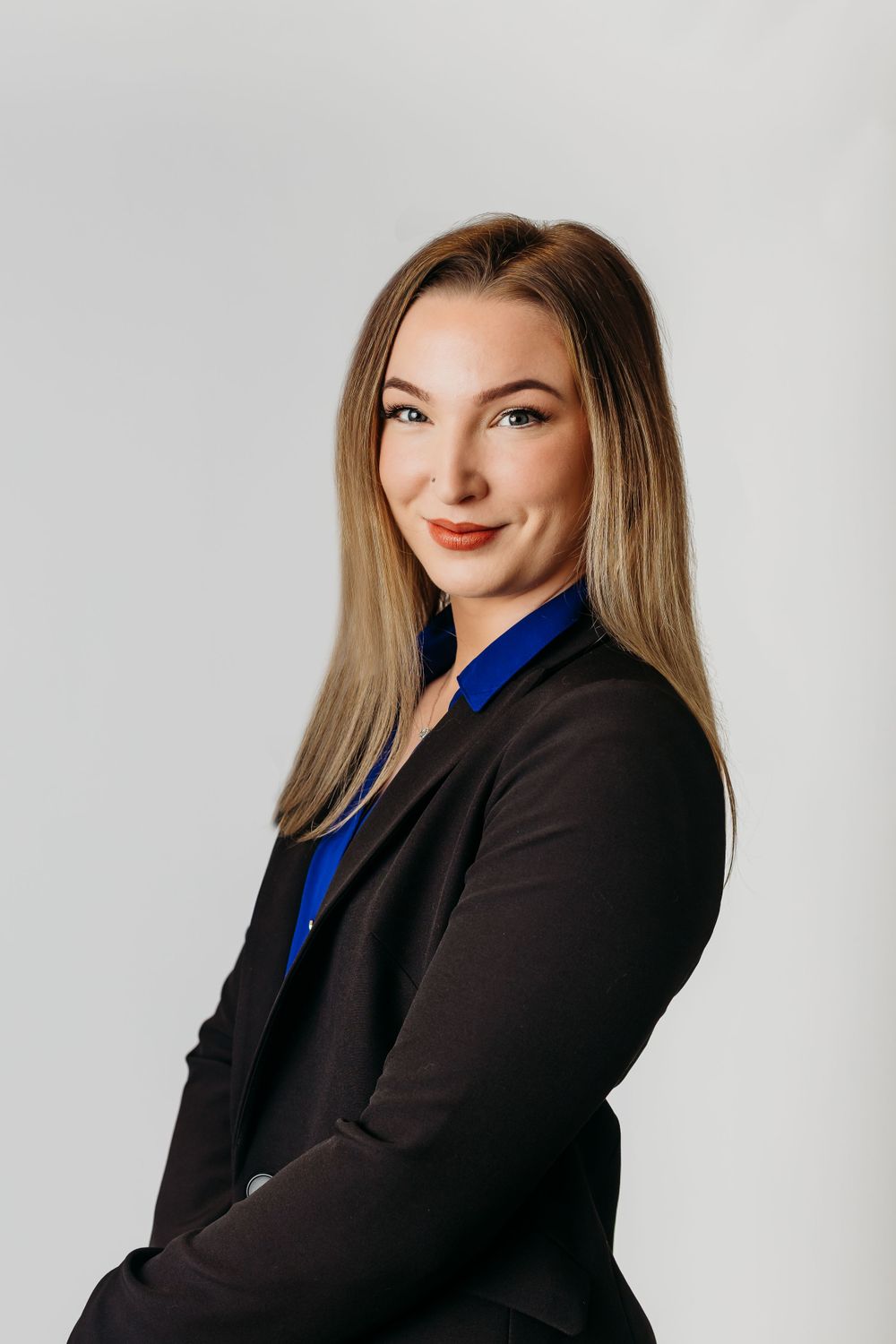 Professional headshot of a business professional in black blazer and blue shirt against grey background.