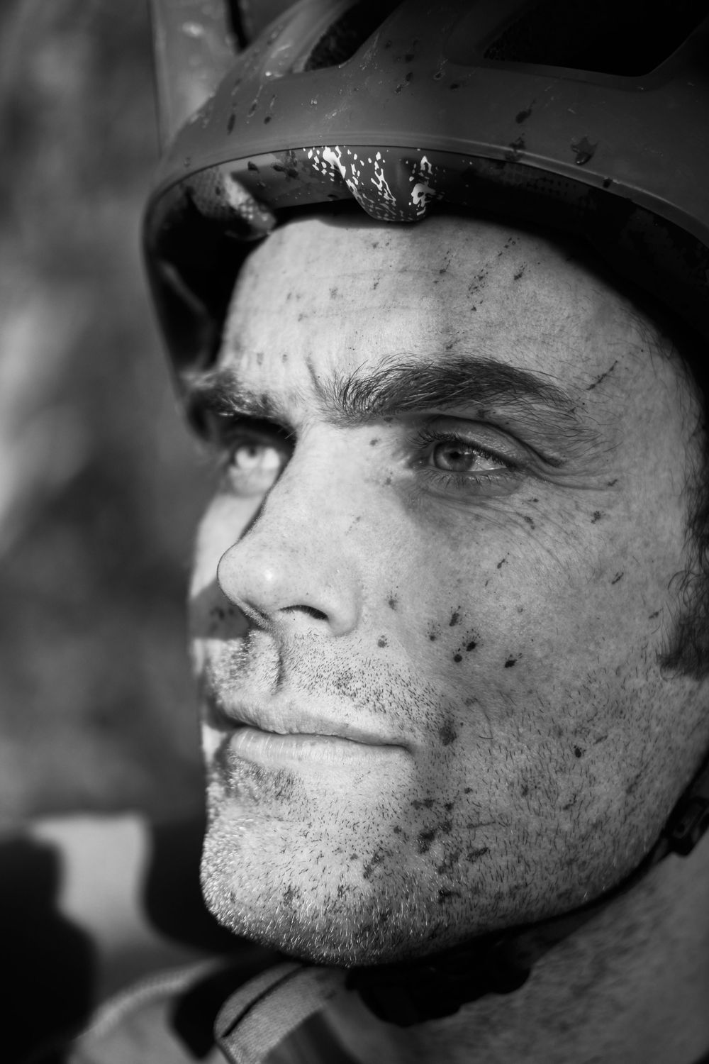 Black and white close up image of a mountain biker's muddy face. Stong beam of light across his face.