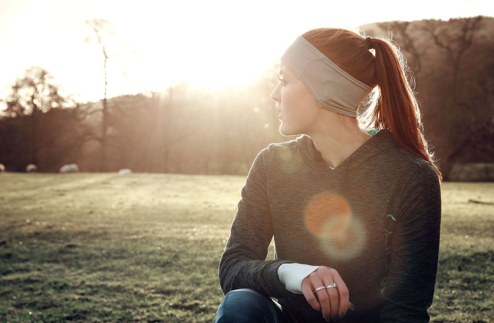 Lifestyle image of a female athlete resting - has headband and ponytail. Dramatic low sunlight, with fields and trees.