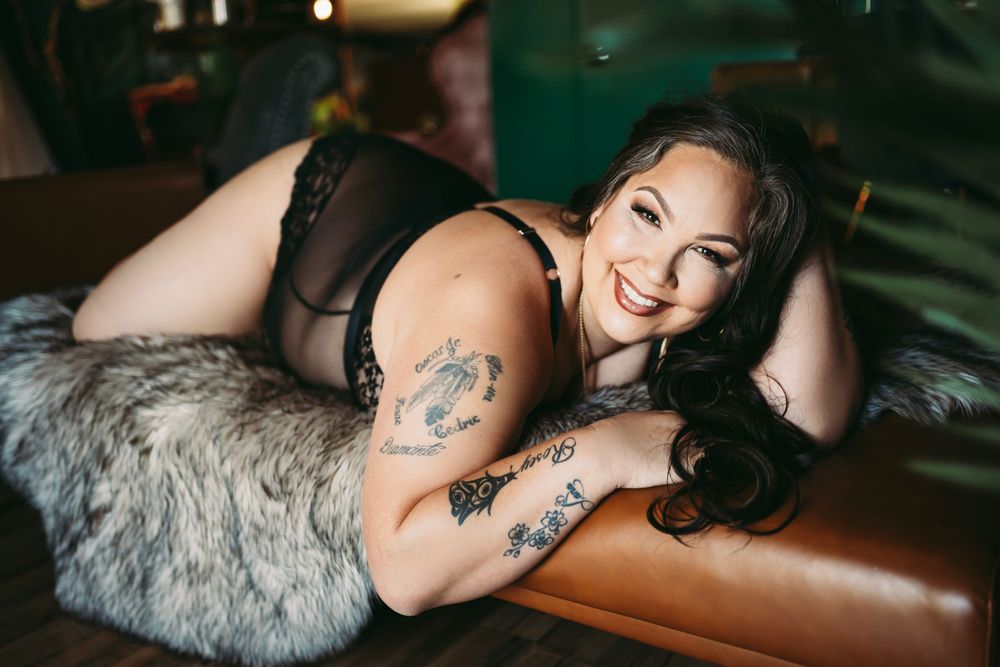 Woman in black lingerie, lying on a leather couch with a cozy and confident expression, highlighting her tattoos and curves.