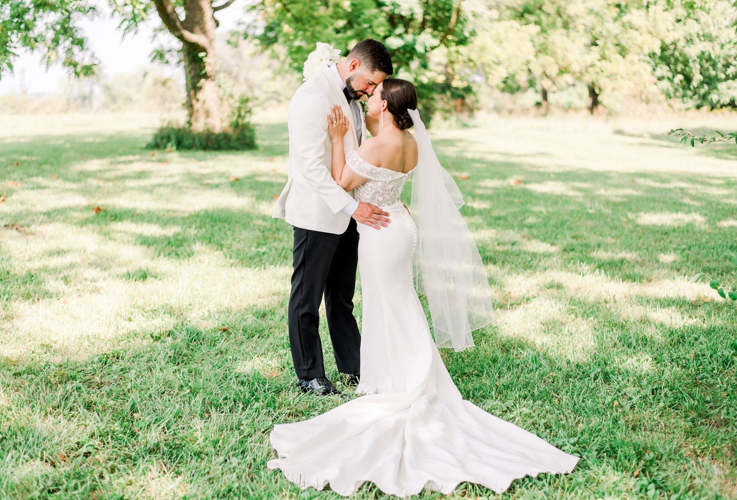 A couple shares an intimate moment on a sunny lawn surrounded by lush greenery and trees.