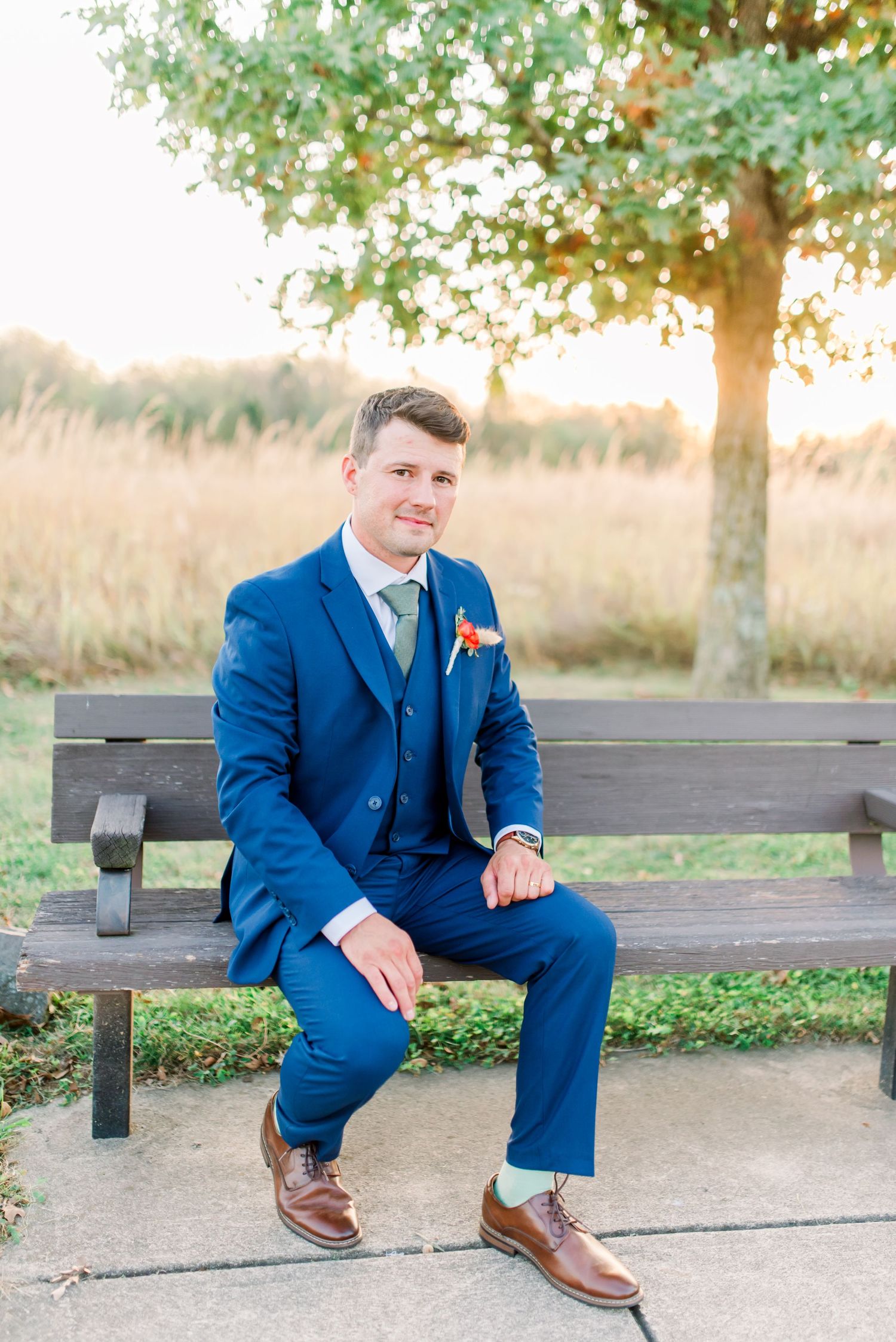 A person in a navy blue suit sits on a wooden bench in a park setting during golden hour.