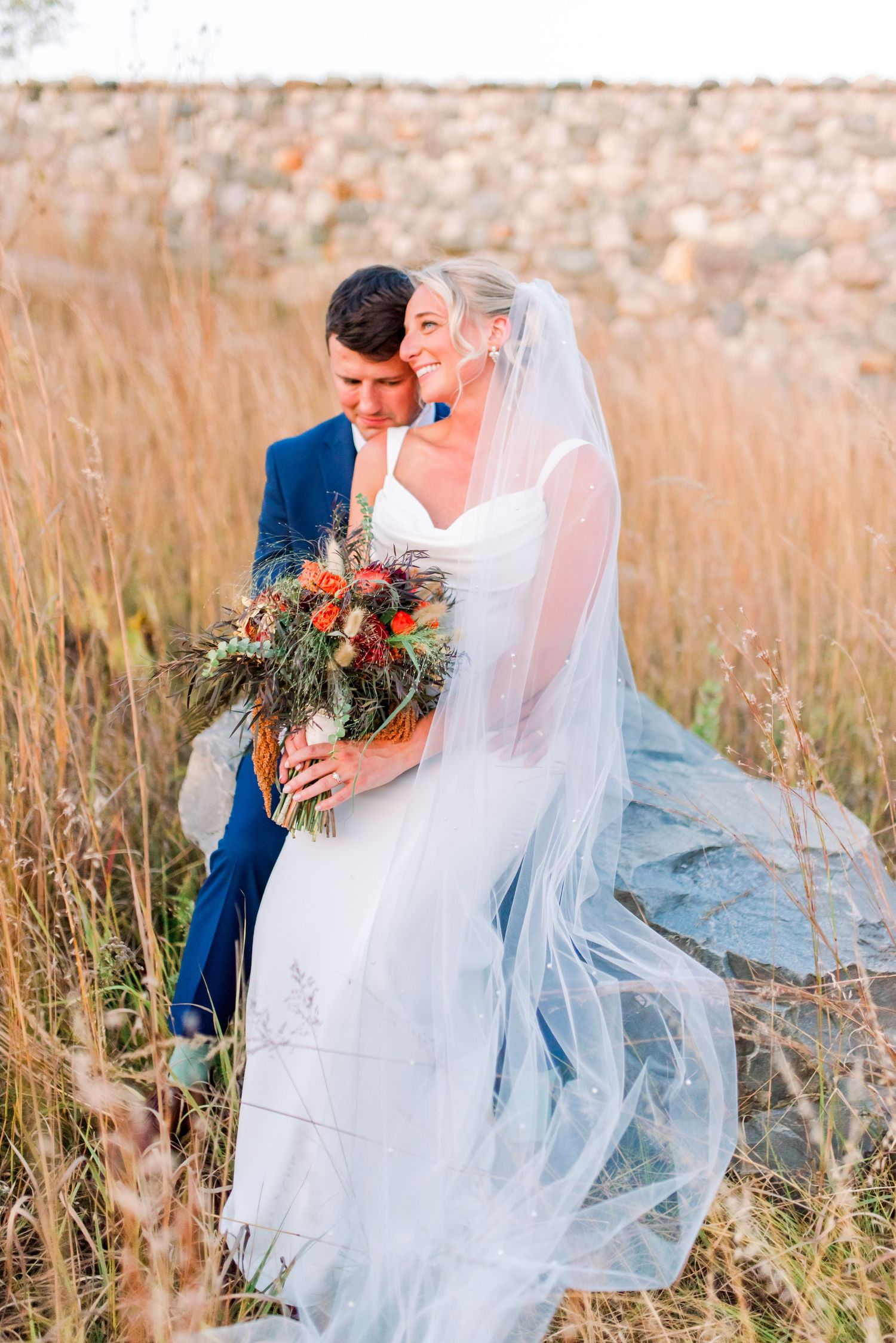 Newlywed couple embraces in a sunlit field of tall grass with flowing veil and floral bouquet.