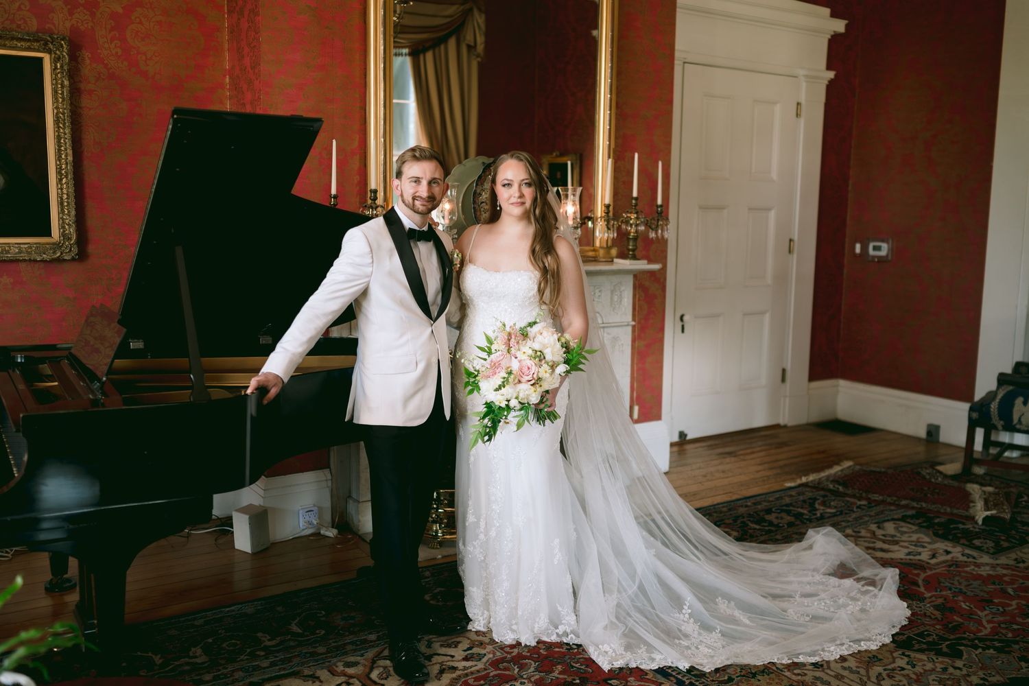 Bride and groom posing elegantly beside a grand piano in a luxurious red-walled wedding venue.
