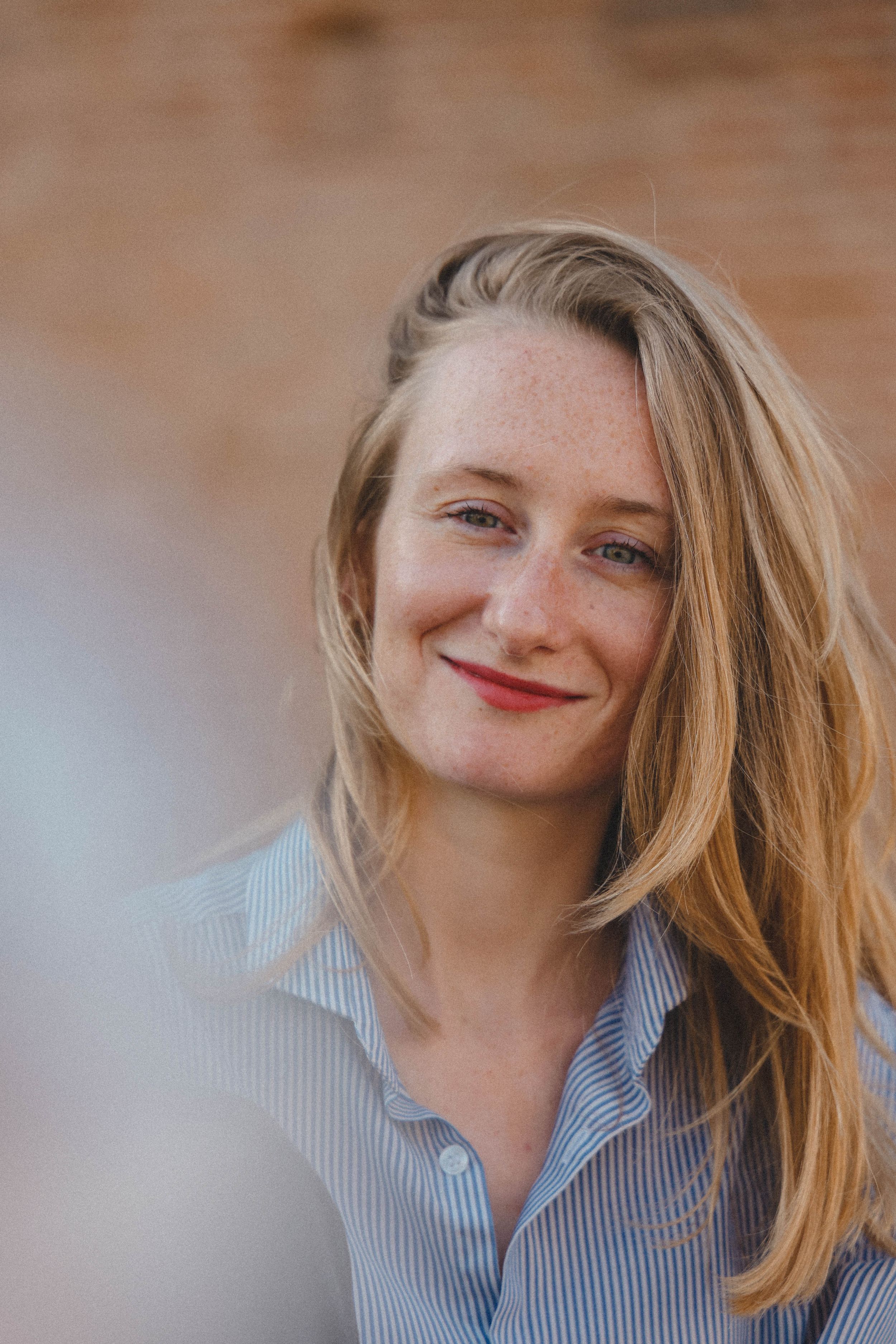 Young blonde woman with freckles and red lips smiling softly in a light blue striped shirt outdoors.