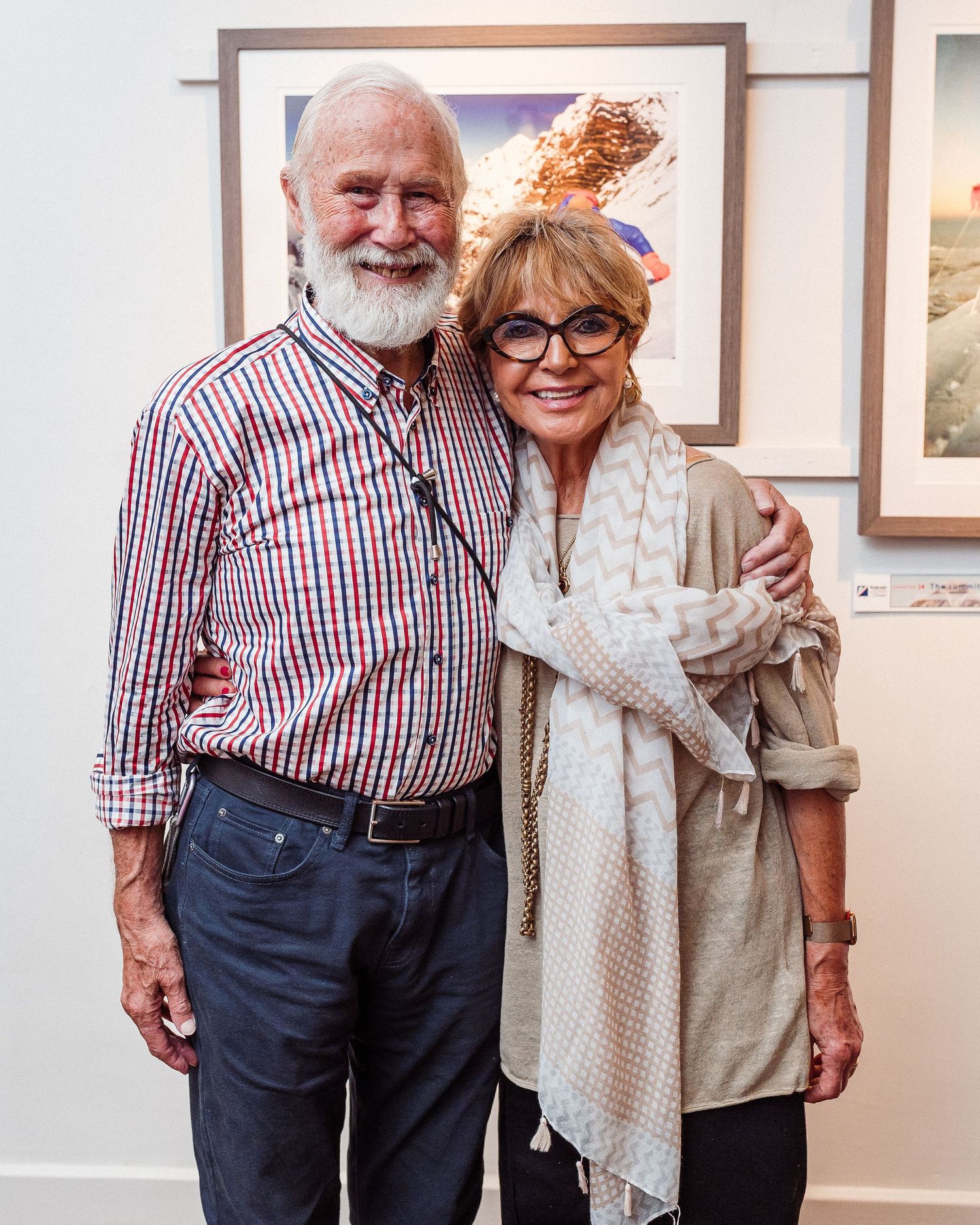 An elderly couple embraces and smiles together while standing in what appears to be an art gallery.