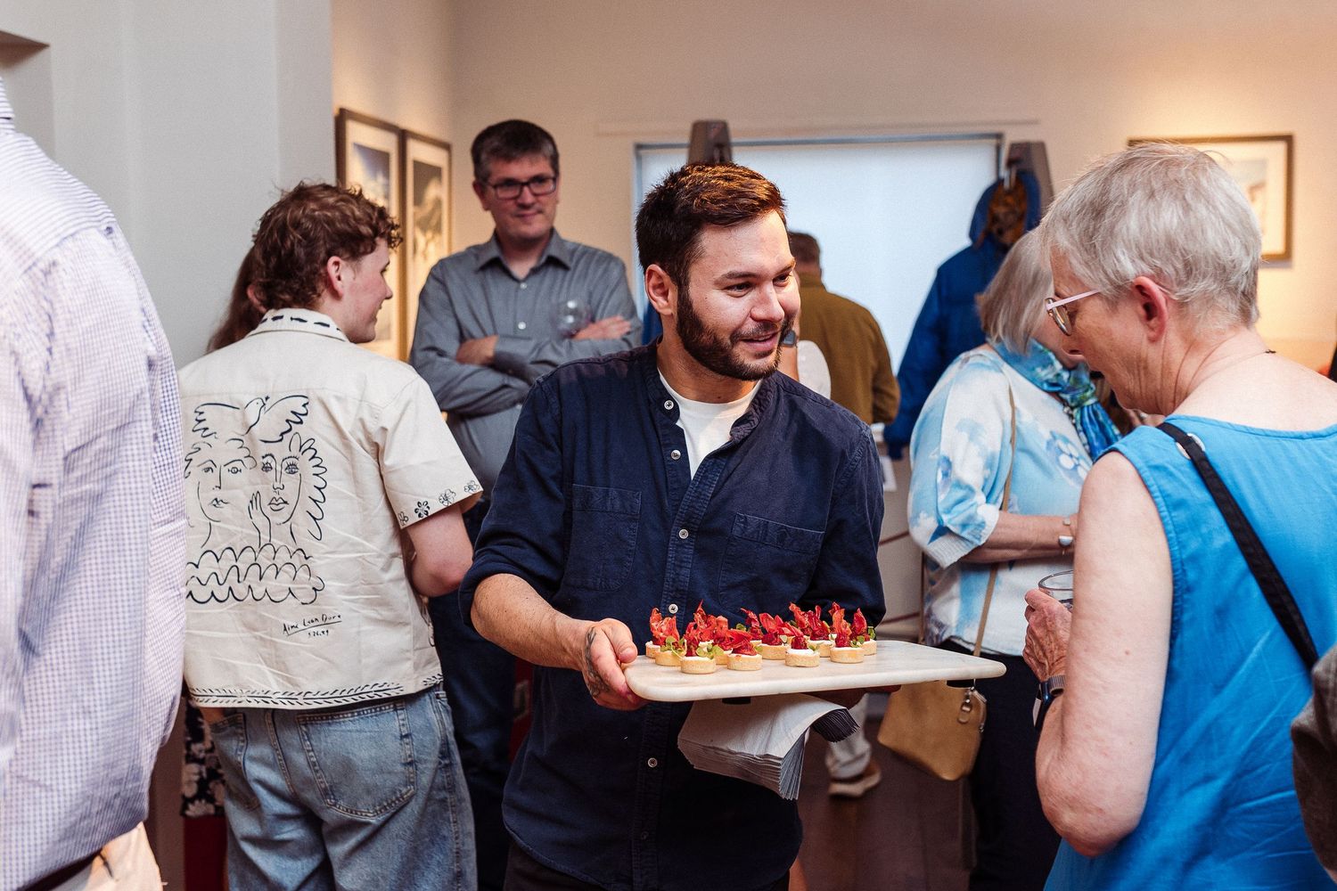 People gather around a table at a social event, talking and sharing food in a casual indoor setting.