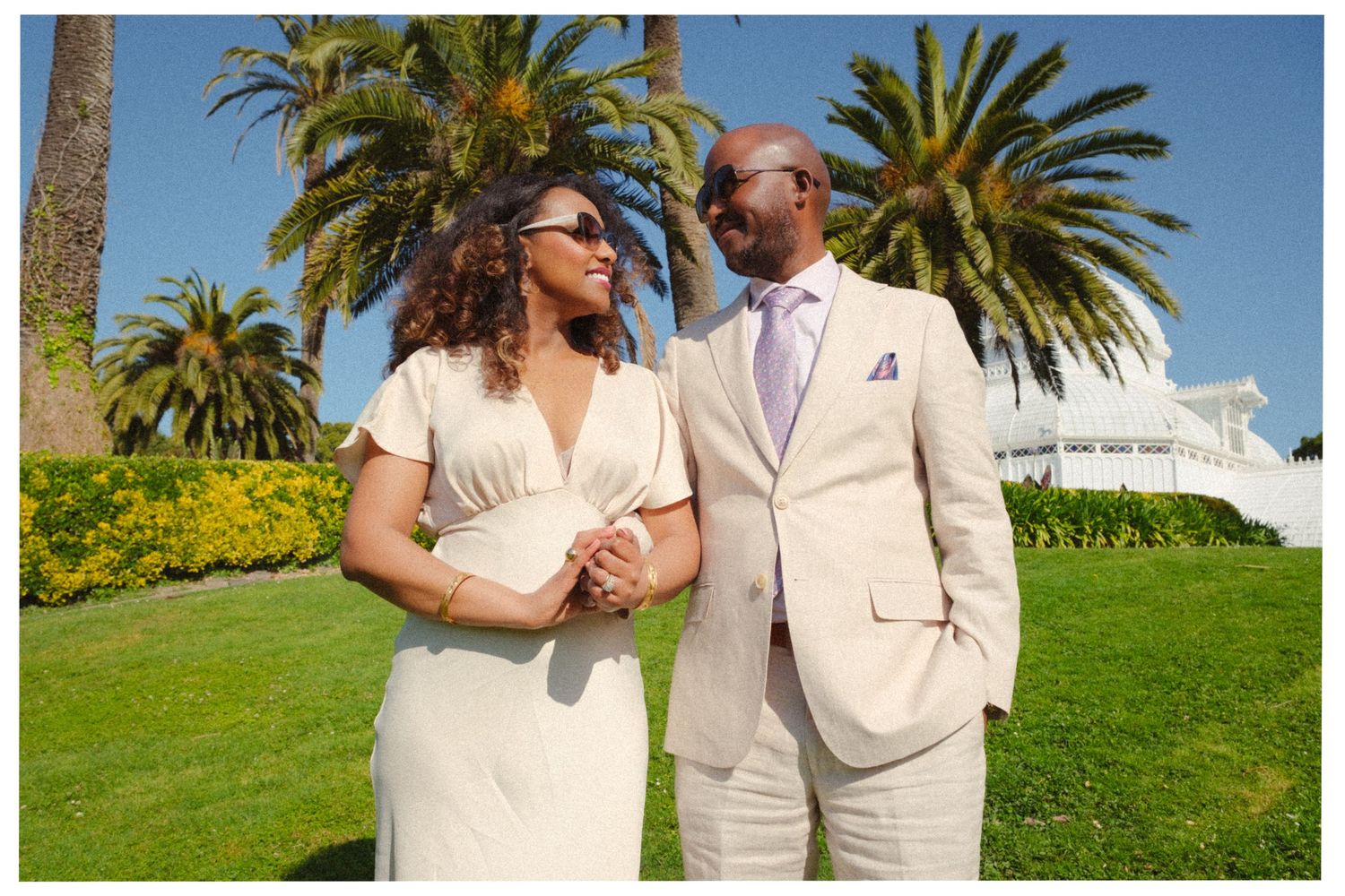 A couple in cream-colored wedding attire walk arm in arm on a lawn with palm trees and a building in the background.