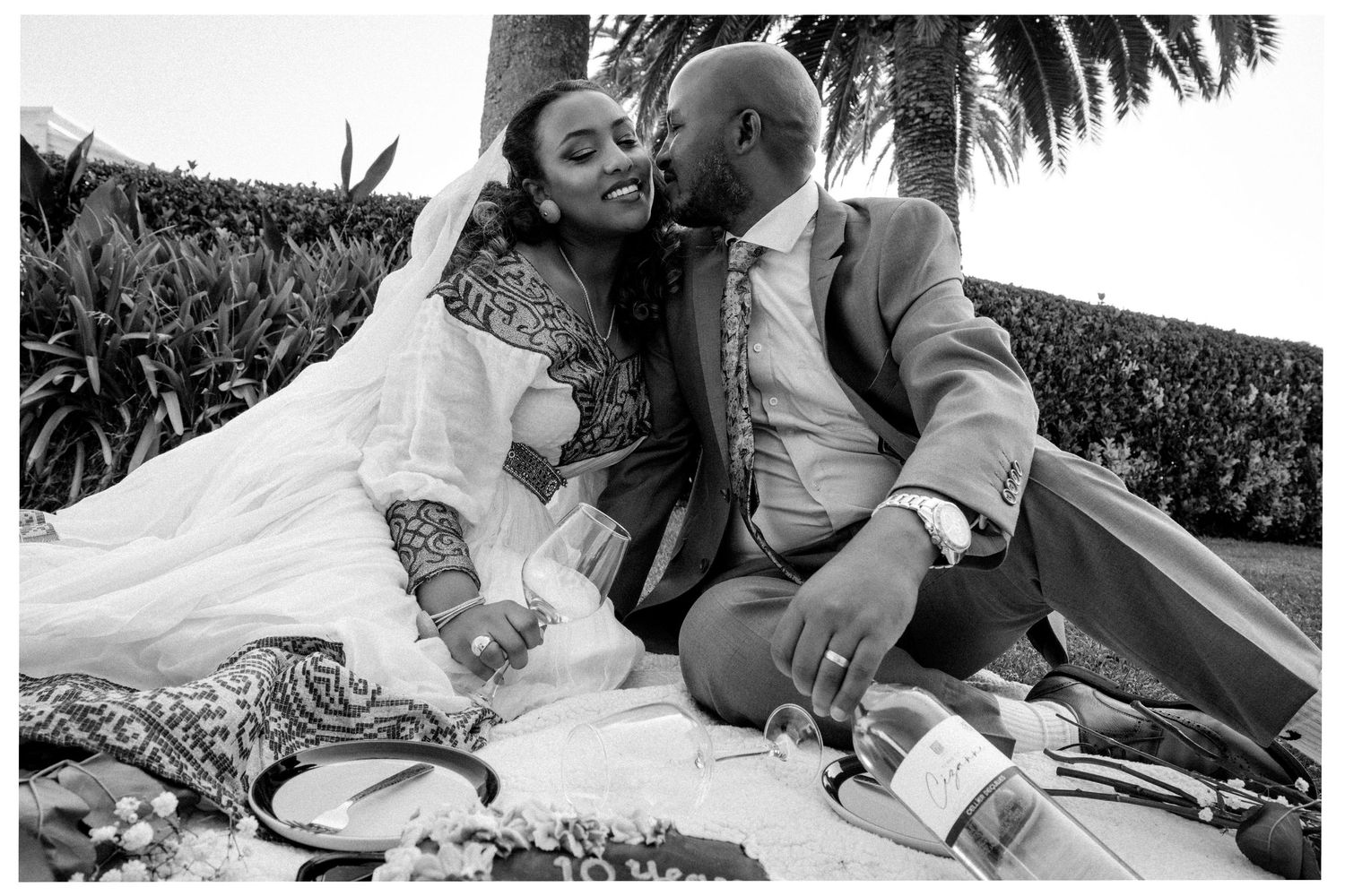 A romantic outdoor picnic moment showing a couple sharing an intimate kiss on a blanket during their wedding day.