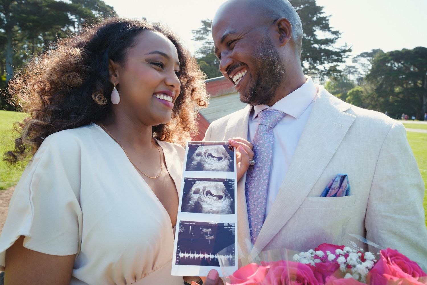 A couple in cream colored outfits share joyful laughter while holding up an ultrasound photo and flowers in a park.