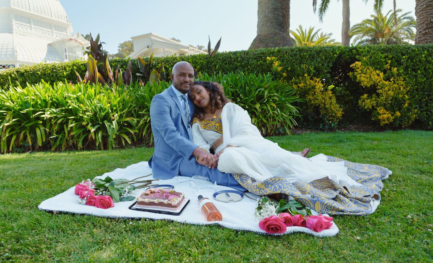 Couple enjoys romantic picnic on a lawn with white blanket, pink flowers and snacks beside tropical garden landscaping.