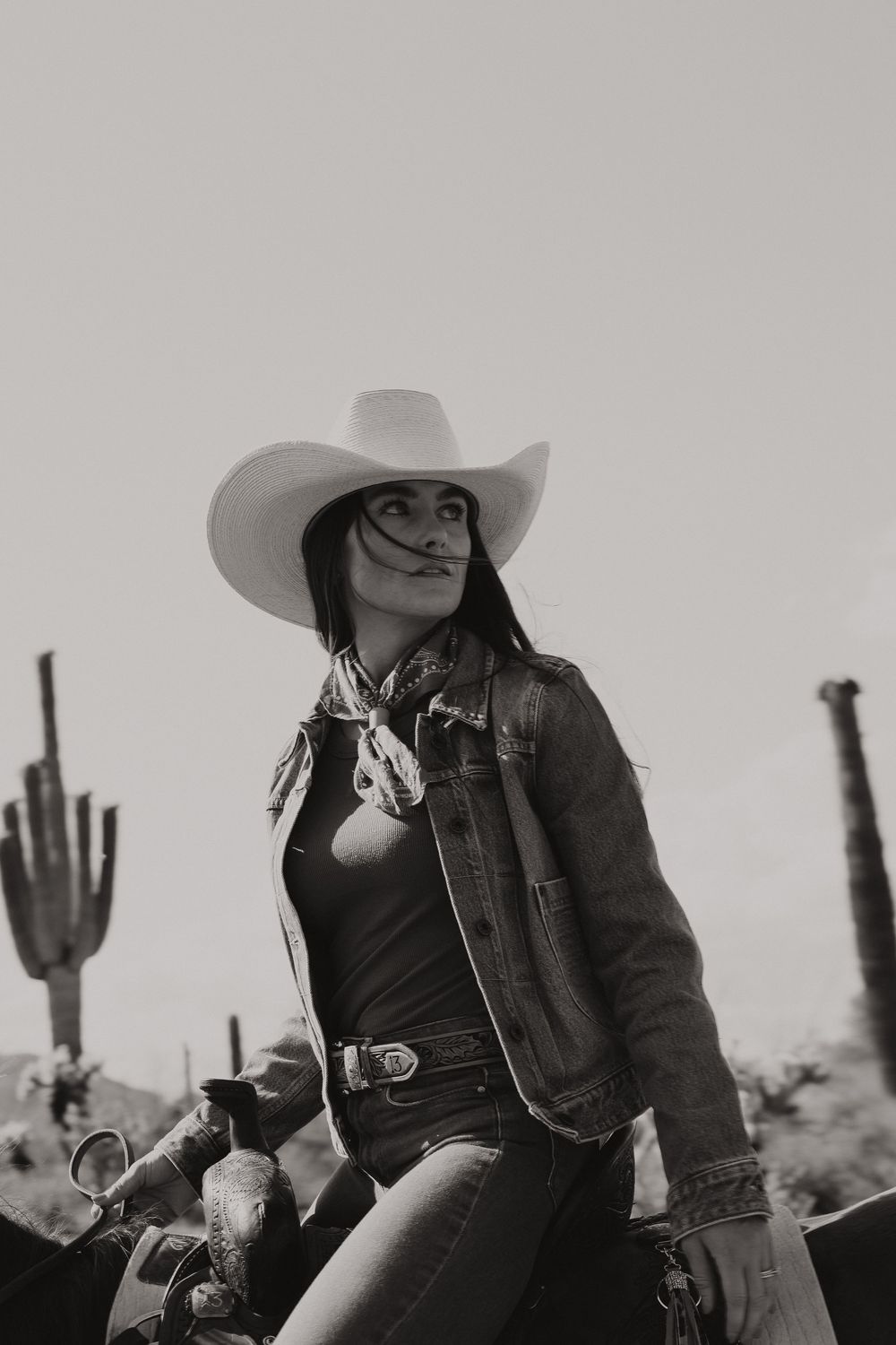 Black and white photo of a cowgirl in a denim jacket and wide brim hat sitting on a motorcycle in a desert landscape.