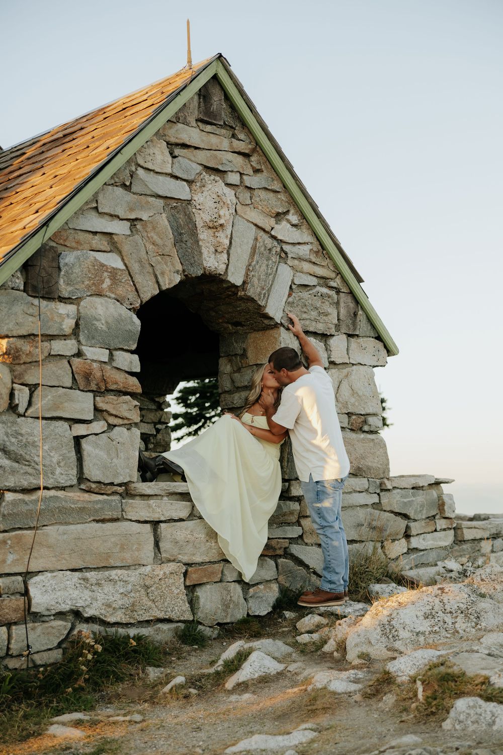 A person in white fabric peeks out from behind a stone shelter on a rocky hilltop at sunset.