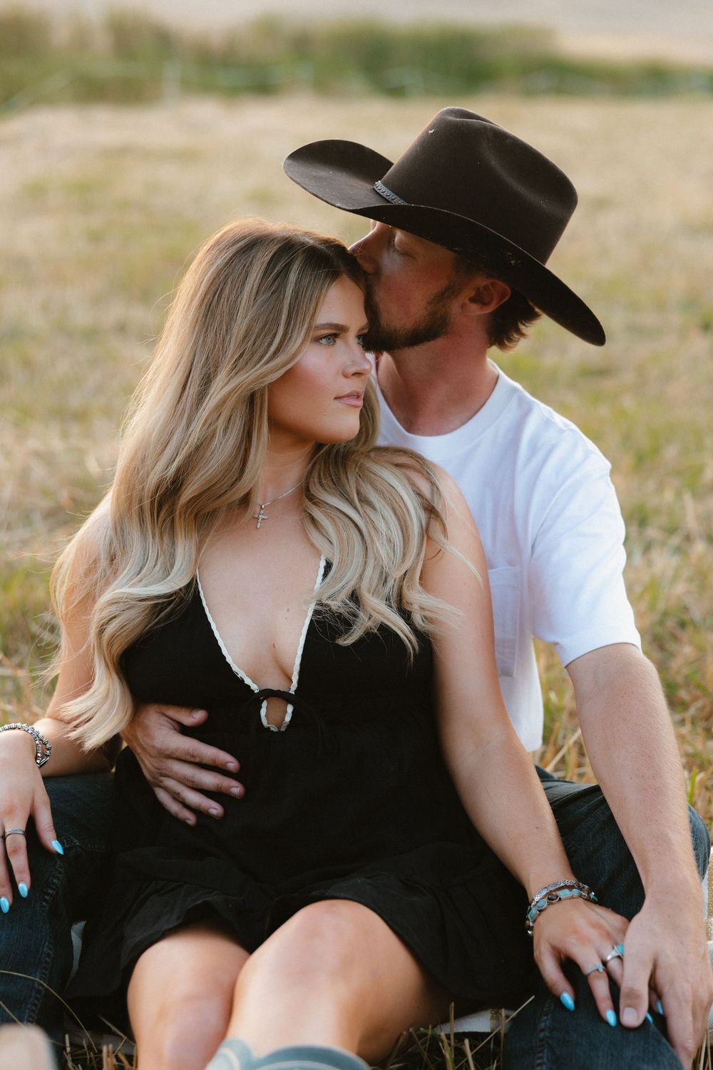 A romantic couple sits in a field, with one wearing a black cowboy hat and white shirt while embracing in an intimate moment.
