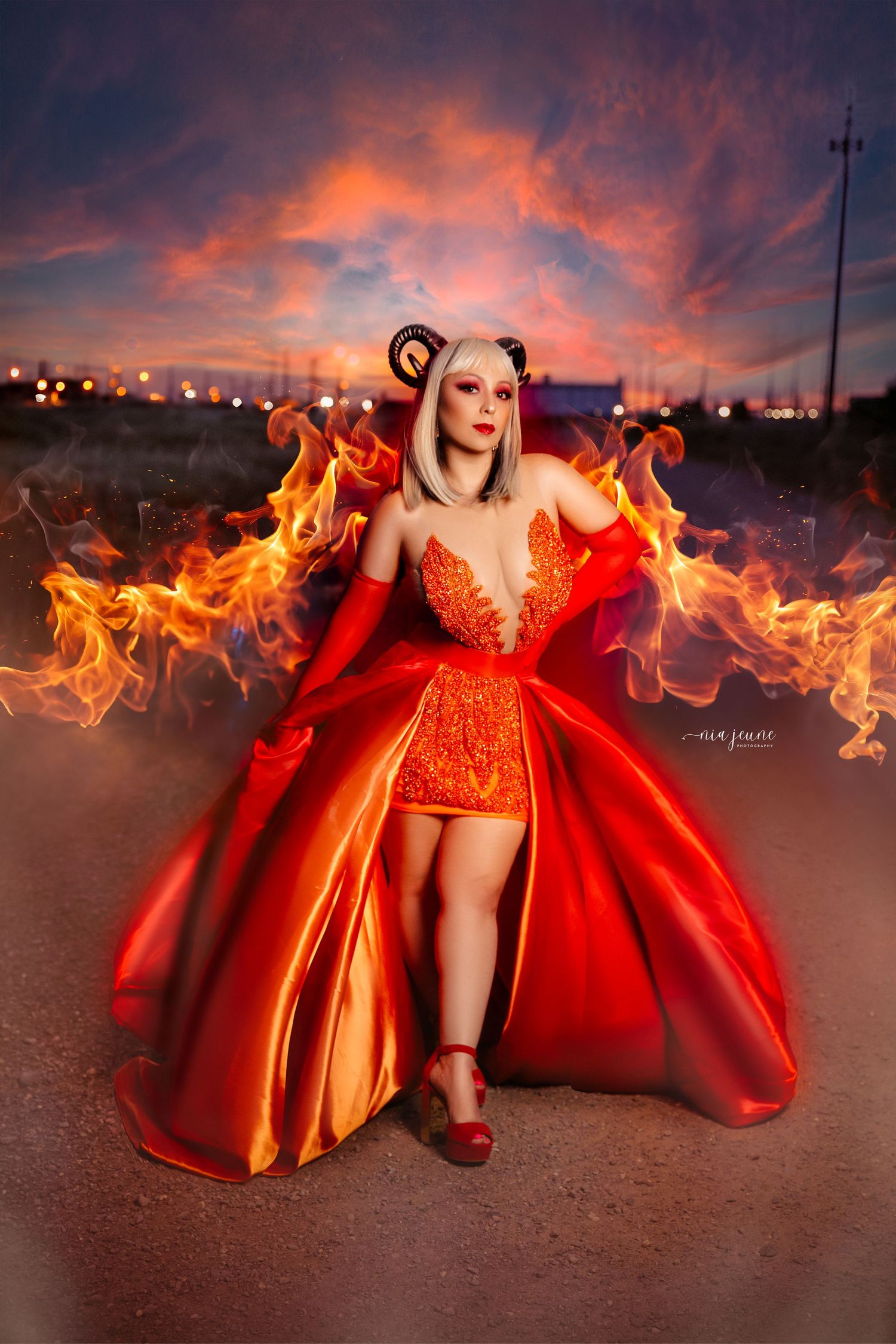 Fantasy portrait with dramatic fire effects around red ballgown against sunset sky.