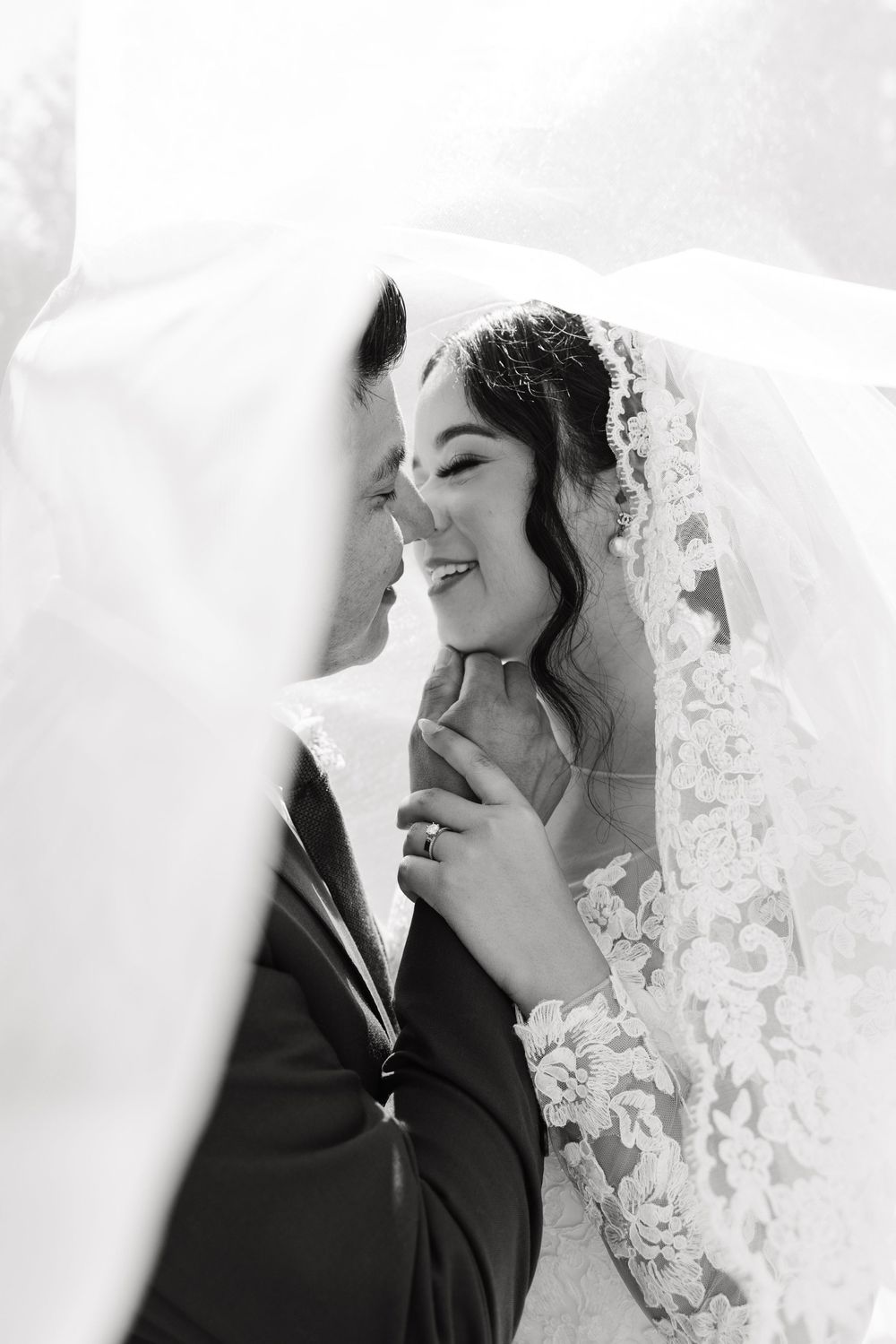 A romantic black and white wedding portrait shows a joyful couple sharing an intimate moment under a delicate lace veil.