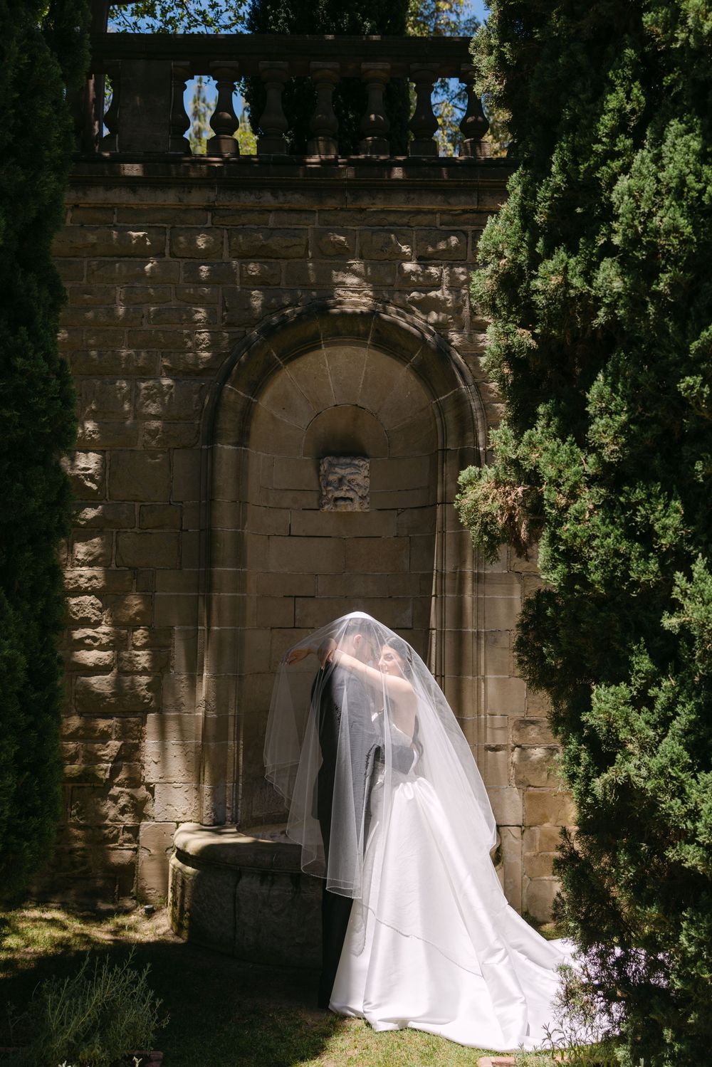 A white wedding gown and veil silhouetted against a stone archway in a garden setting, casting dramatic shadows.