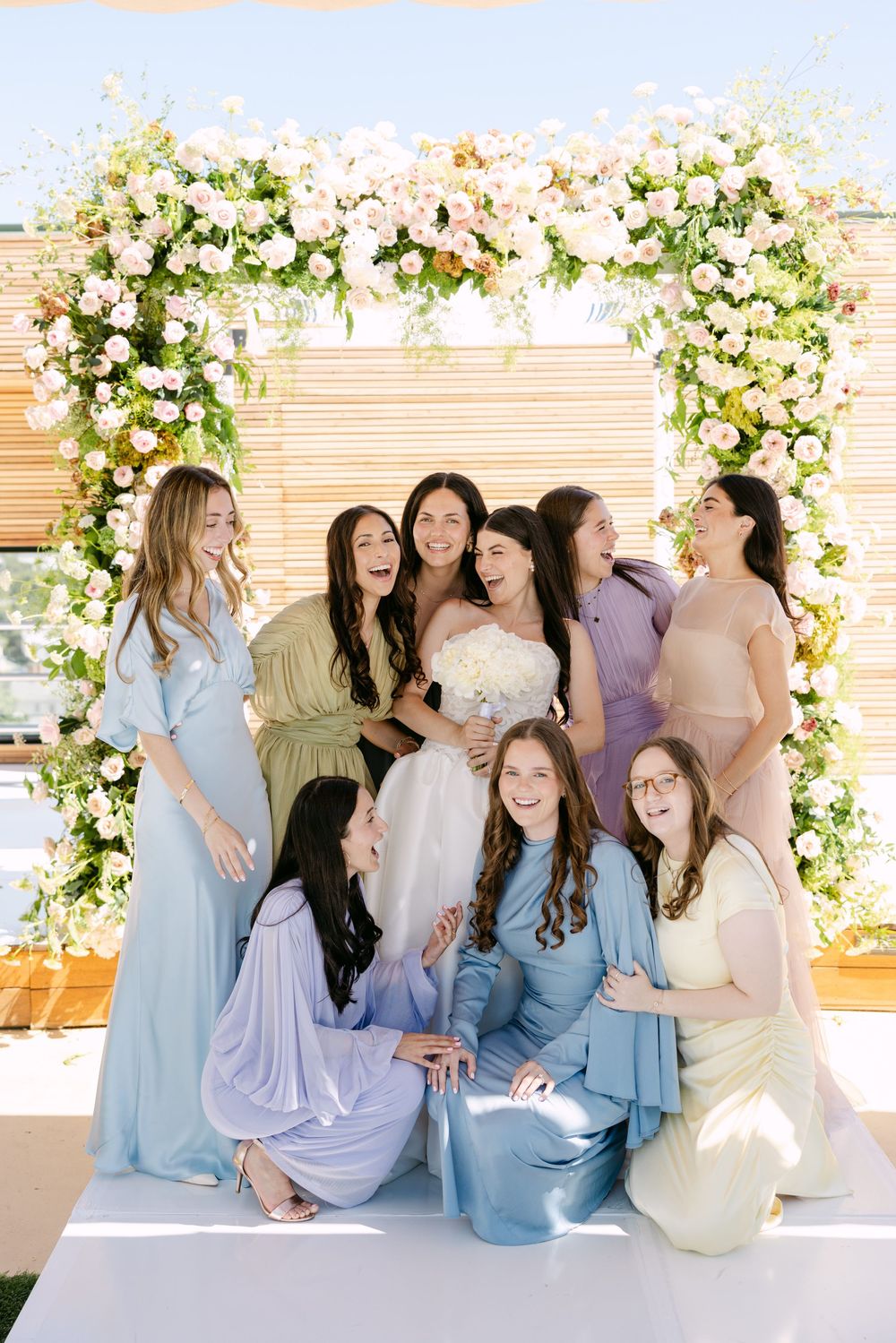 Bridesmaids wearing pastel blue dresses pose together in front of a floral arch with white and peach blooms.