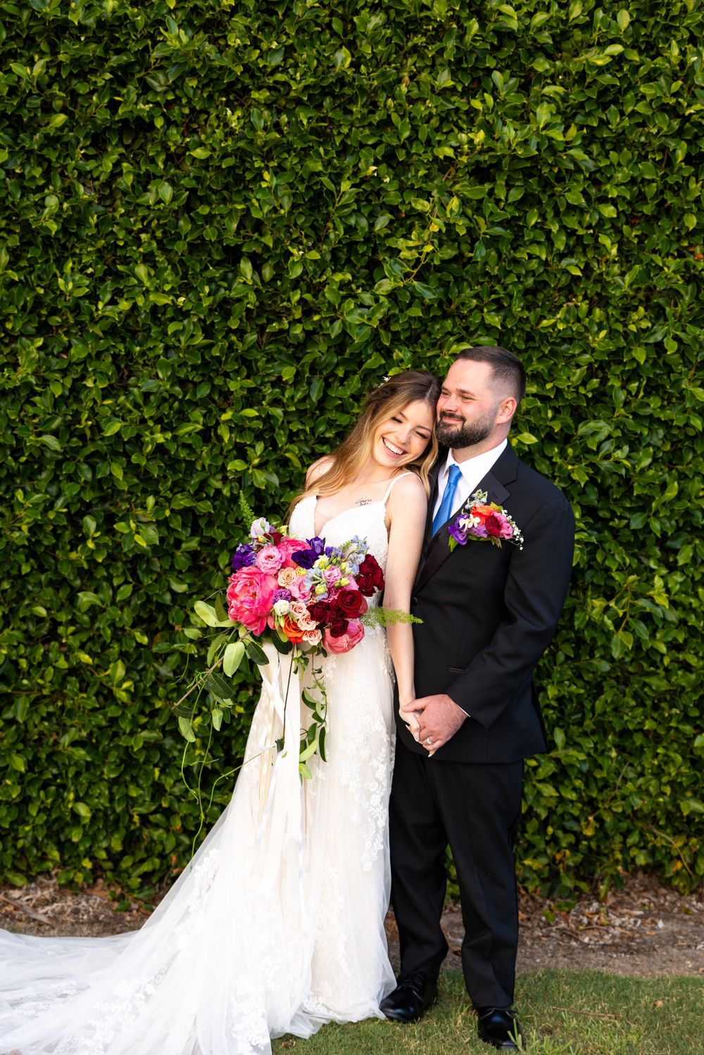 A couple poses against a lush green hedge wall with vibrant pink and purple bouquet floral arrangements.