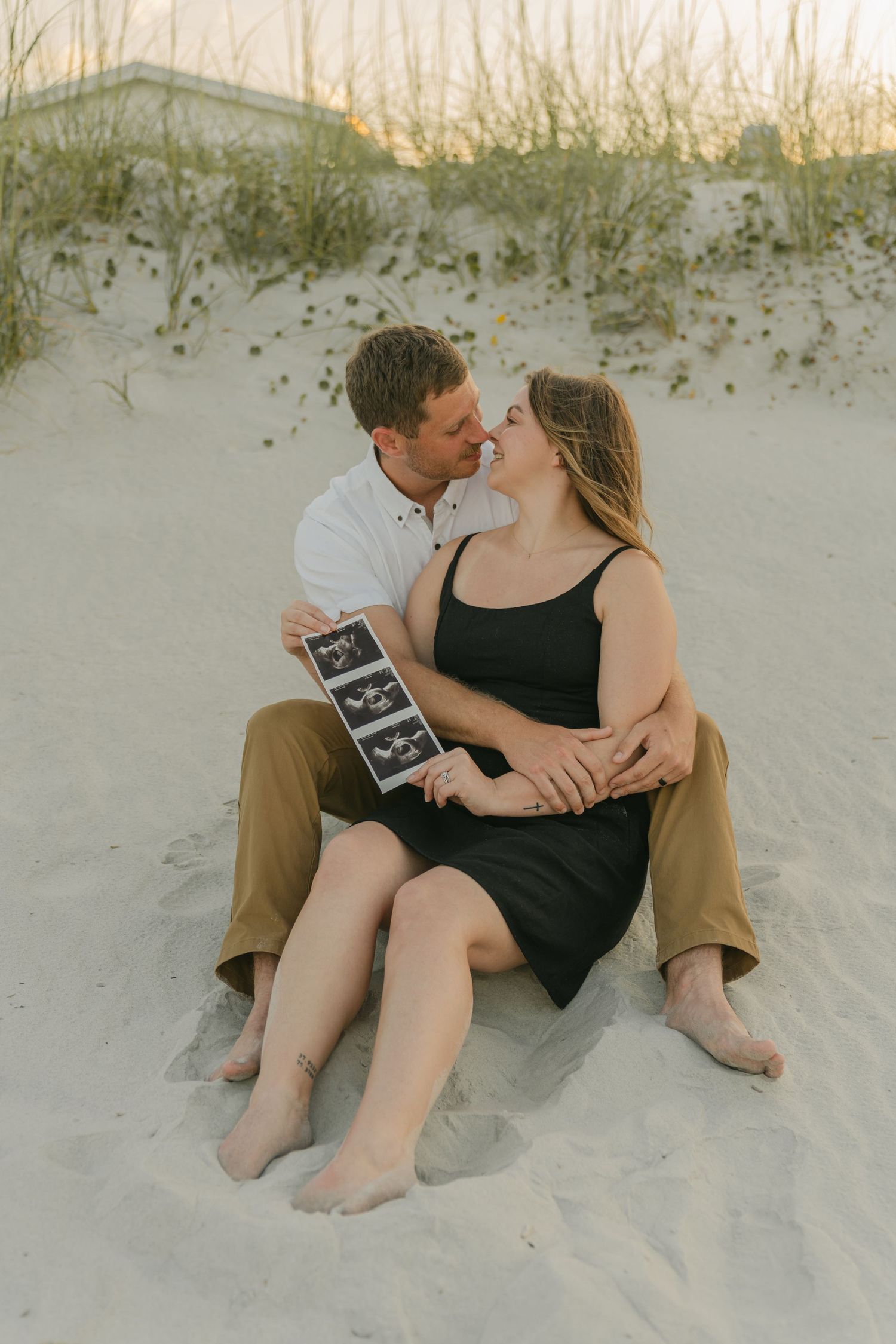 Couple sitting intimately on sandy beach dunes at sunset holding signs.