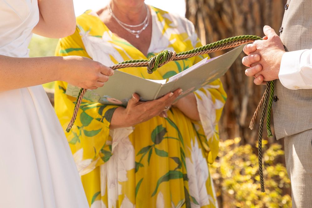 Officiant in yellow floral dress reads from book during outdoor wedding handfasting ceremony.