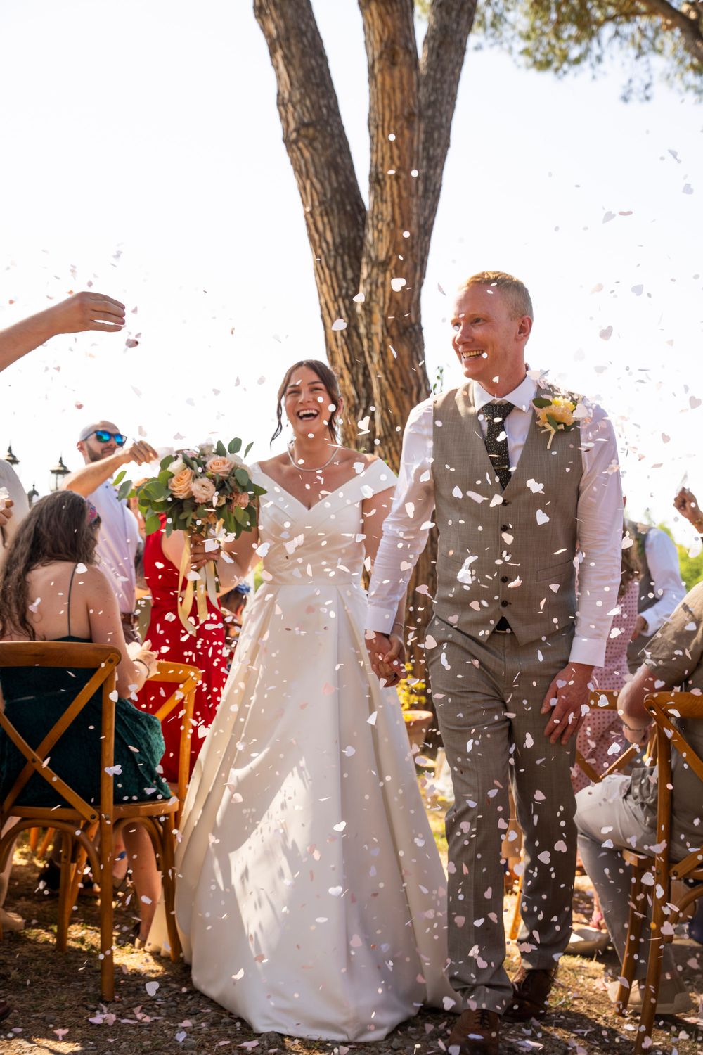 Bride and groom walk down the aisle as guests toss white confetti outdoors under sunny skies.