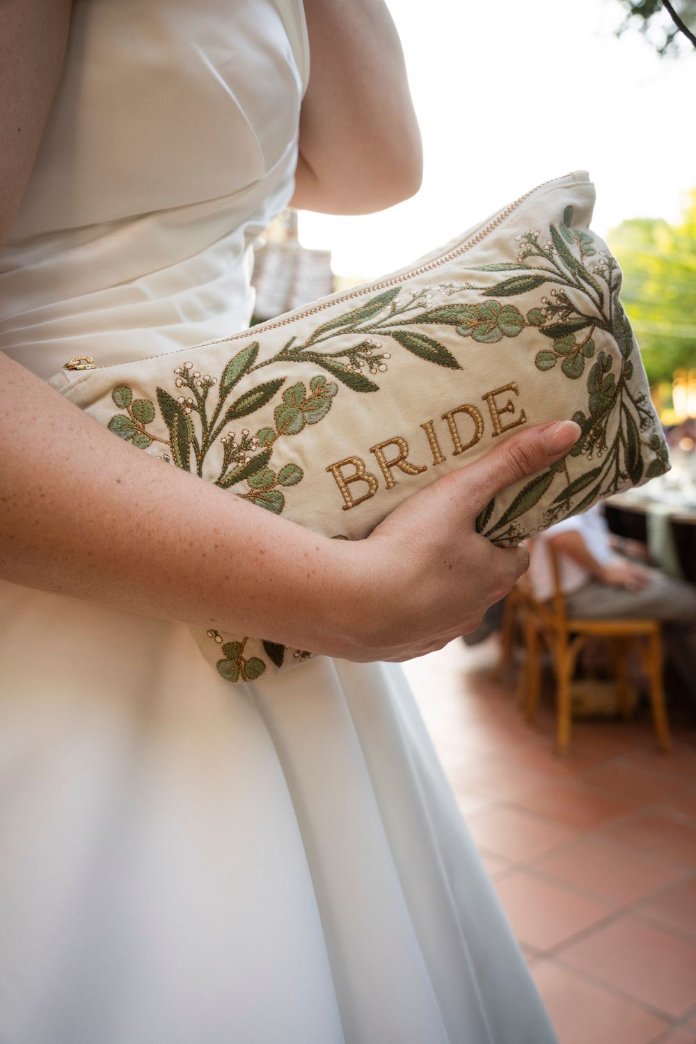 Bride holds an embroidered floral clutch bag with the word BRIDE on her wedding day.